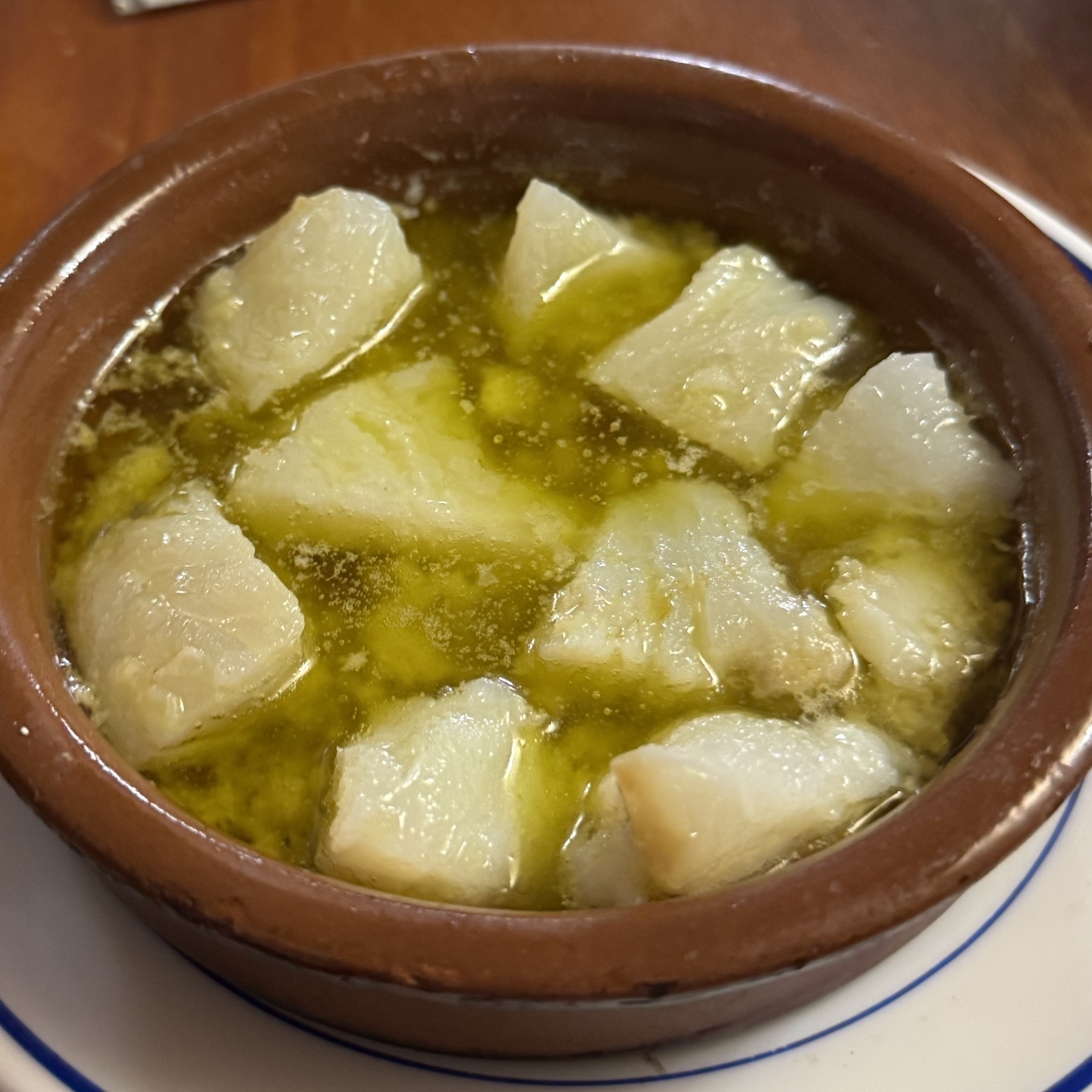Clay dish containing chunks of cod simmering in olive oil, with the fish appearing tender and slightly translucent.