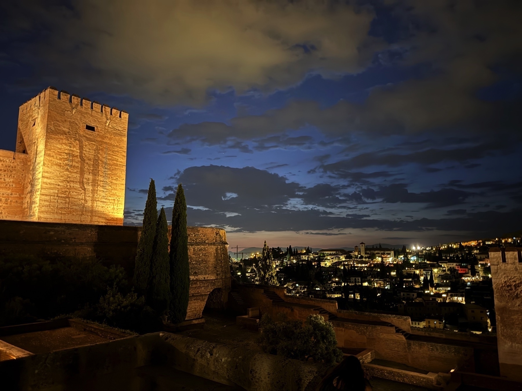 View of the Torre de la Vela at the Alhambra lit against a deep blue twilight sky, with the illuminated Albaicín neighborhood sprawling across the valley in the background under dramatic clouds.