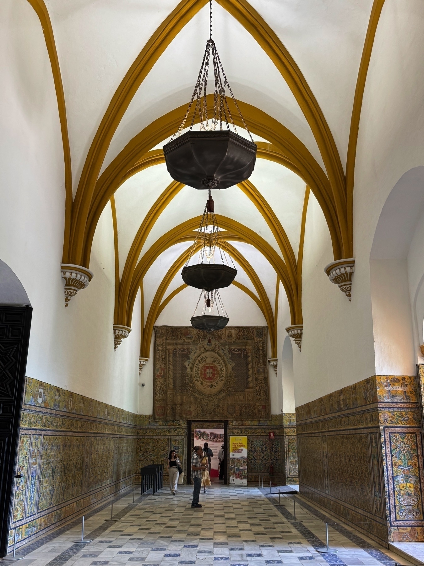 The Gothic Hall with high ribbed vaults painted in gold and black, large iron lanterns suspended from the ceiling, and walls clad in colorful azulejo tile. A large tapestry bearing a heraldic emblem hangs above a central doorway.
