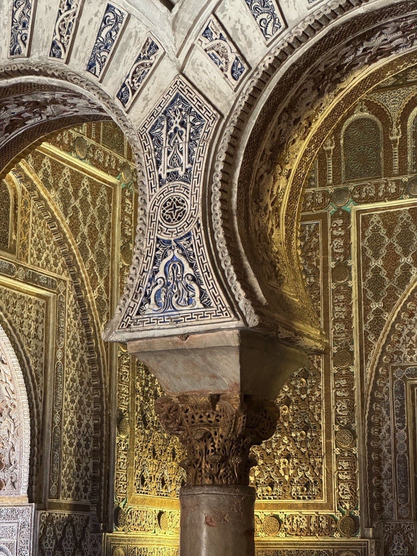 A polylobed arch supported by a decorated column in the Hall of Ambassadors. The surface is covered in blue-and-white ceramic inlay and dense Mudéjar stucco work with floral and geometric motifs.