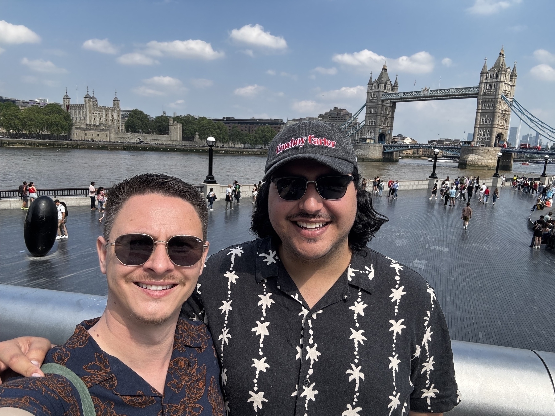 Two smiling people wearing sunglasses pose for a selfie on a sunny London day with Tower Bridge and the Tower of London in the background.
