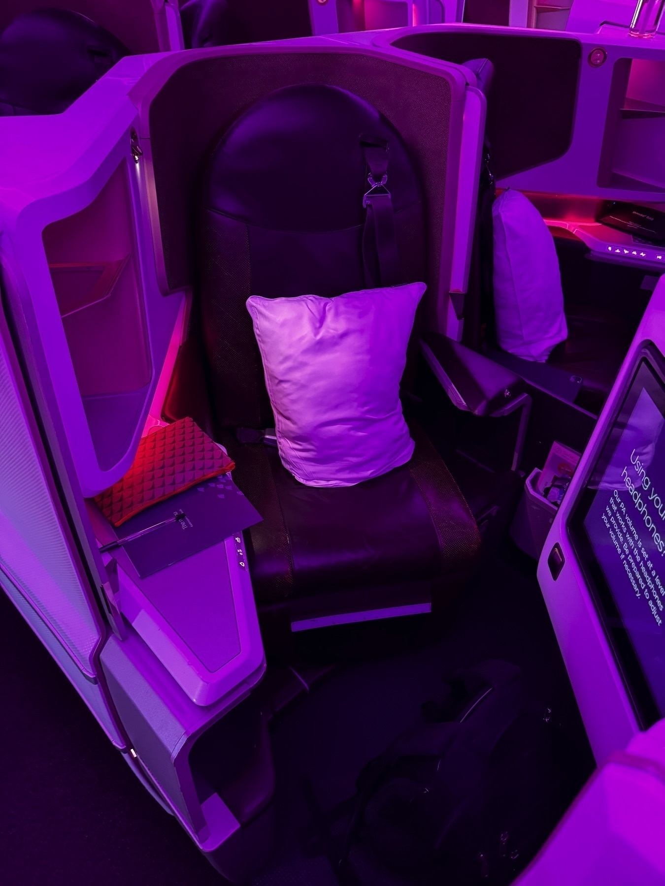 Empty Virgin Atlantic Upper Class seat prepared for boarding, with a leather seat, pillow, and purple mood lighting. The seat is surrounded by storage areas and a folded tray table, emphasizing its spacious and private setup.
