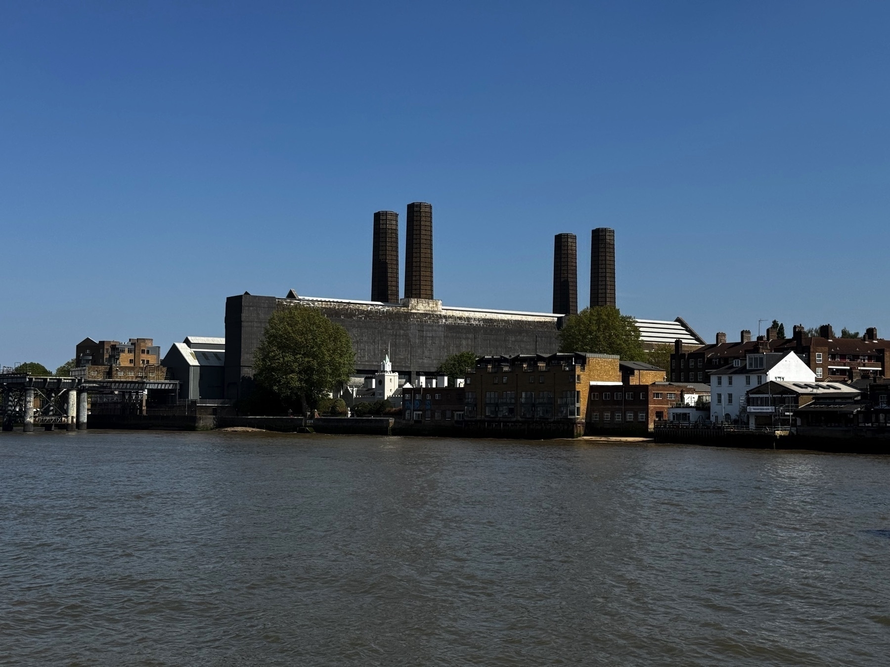 A striking view of a riverside industrial complex with four tall chimneys, likely the former Lots Road Power Station. In front, smaller brick and white-painted buildings sit along the waterfront, forming a contrast with the industrial structure.
