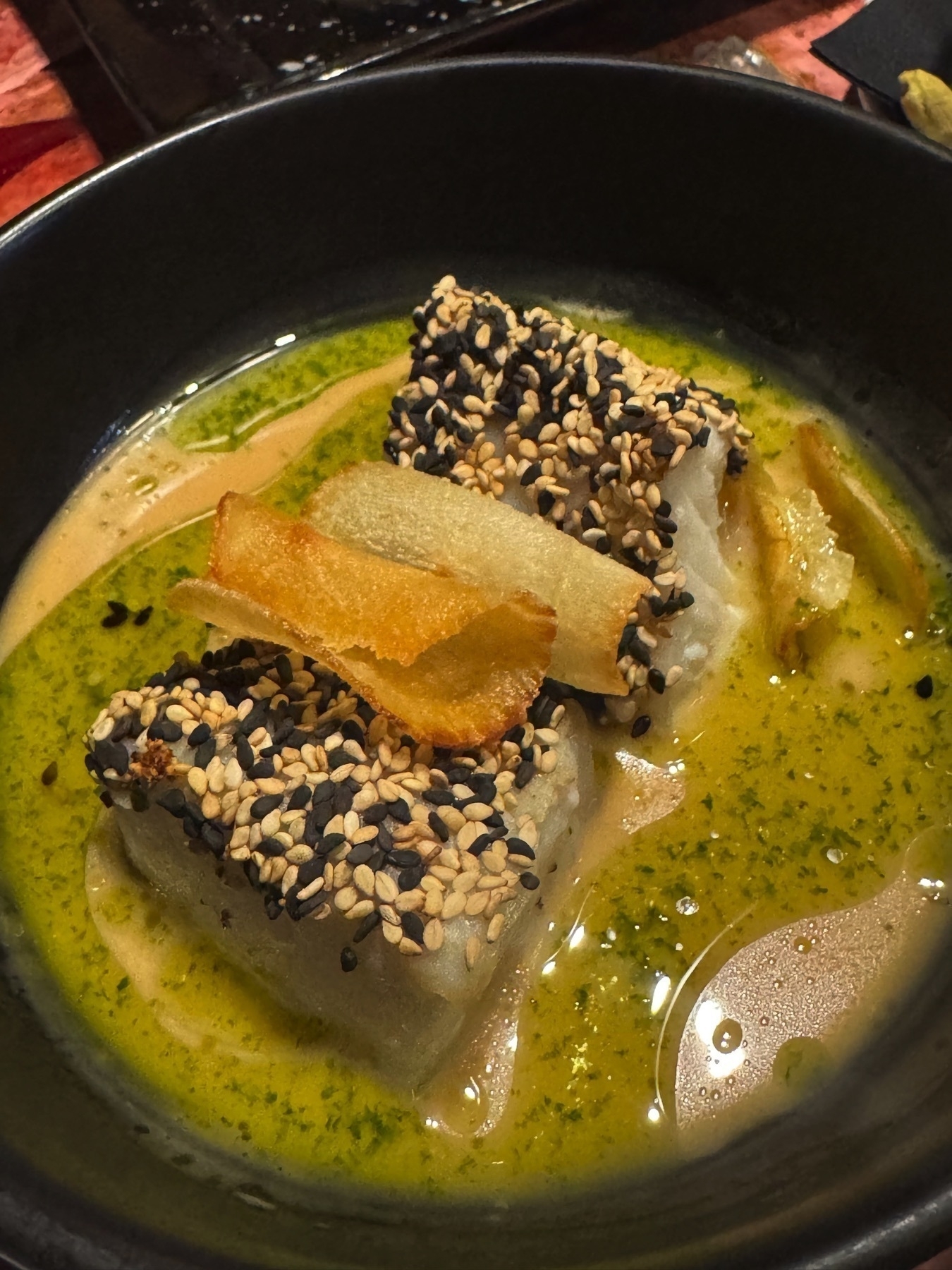 Two pieces of white fish crusted with black and white sesame seeds are served in a shallow green sauce and topped with thin crisps. The glossy broth-like sauce surrounds the fish, adding richness and contrast to the crunchy sesame coating.