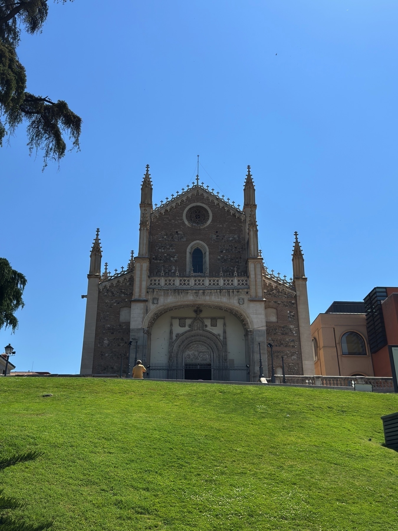 View of San Jerónimo el Real, a Gothic church behind the Museo del Prado. The church’s ornate facade with pointed arches and decorative pinnacles stands out against a deep blue sky. The building is situated atop a grassy hill, where a lone visitor in yellow sits facing the structure, emphasizing its scale and serenity.