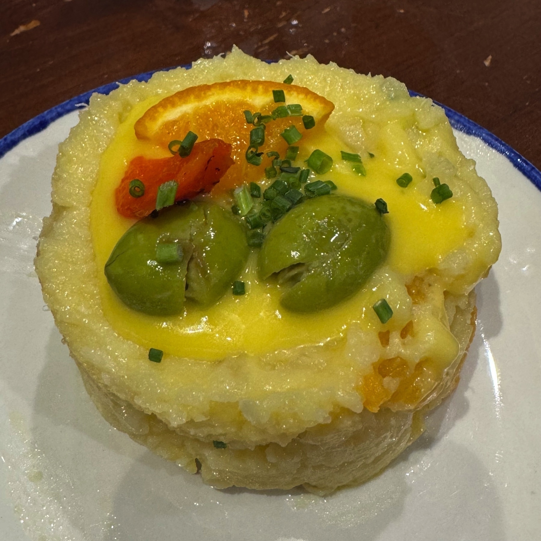 Small round dish of Ensalada Malagueña, made of mashed potatoes topped with bright green olives, a slice of orange, a sliver of roasted red pepper, and a drizzle of vibrant yellow sauce, garnished with chopped chives.