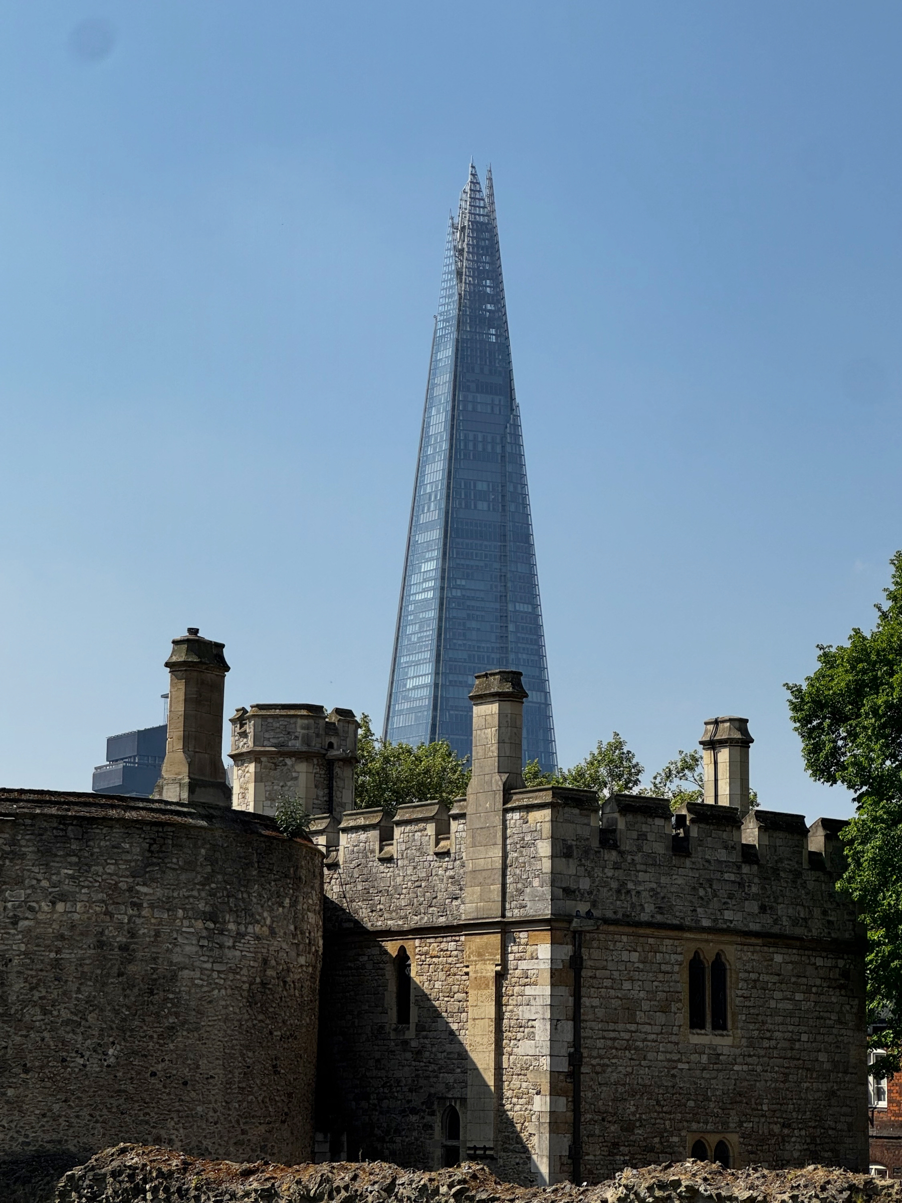 Rugged stone towers and battlements stand in the foreground, their crenellations sharply defined in sunlight, while the Shard rises sleek and reflective behind them against a pale blue sky.