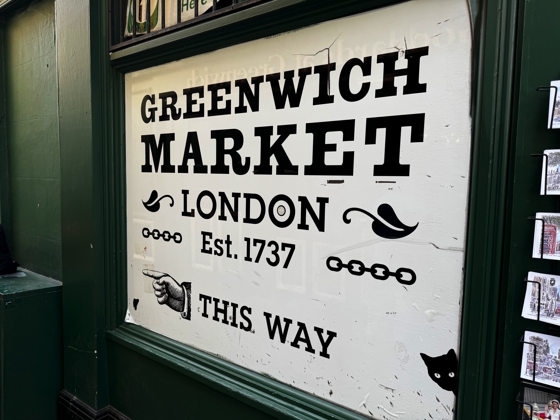 A weathered sign on a white background reads “Greenwich Market London Est. 1737 This Way,” adorned with vintage-style typography, scrollwork, a pointing hand, and a black cat peeking from the corner. The sign is mounted in a dark green frame on an exterior wall, hinting at the historic and quirky charm of the nearby market.