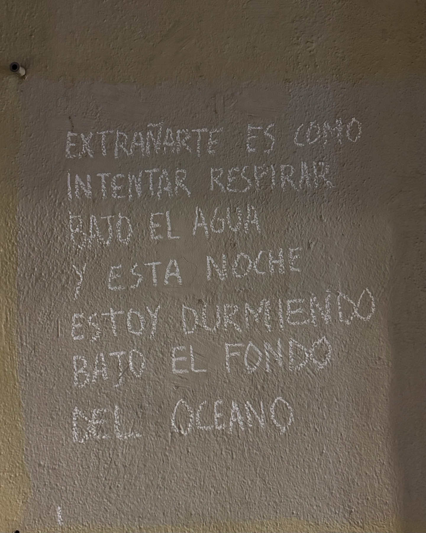 Spanish poem handwritten in white chalk on a rough beige wall in Málaga, reading: “Extrañarte es como intentar respirar bajo el agua y esta noche estoy durmiendo bajo el fondo del océano,” expressing intense longing and emotional isolation.