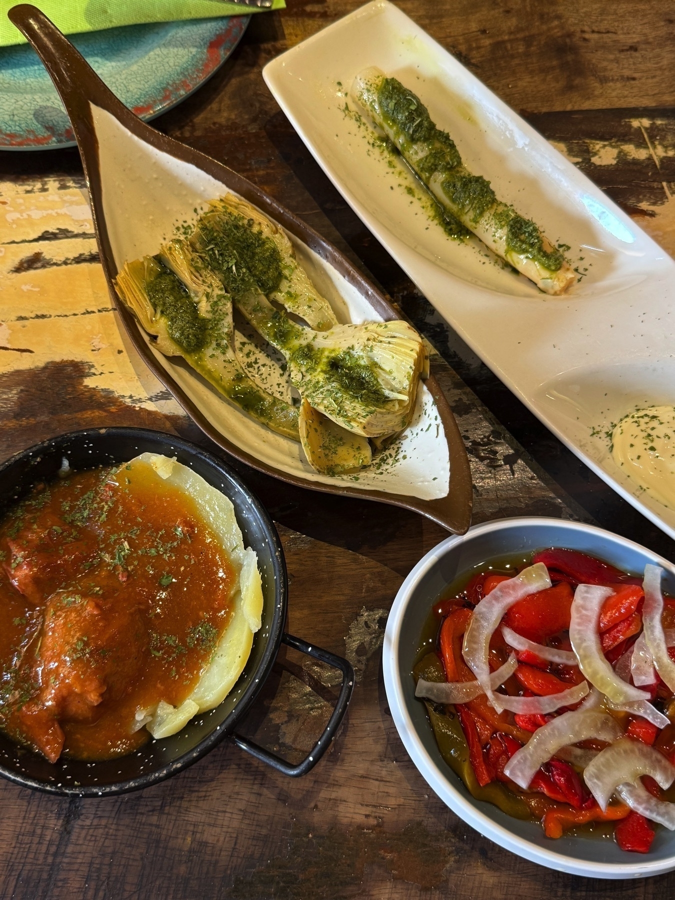 Eclectic spread of Spanish tapas on rustic dishware, including grilled artichokes with herbs, piquillo peppers with onions in olive oil, and a potato and tomato dish, offering a colorful and inviting sampler.