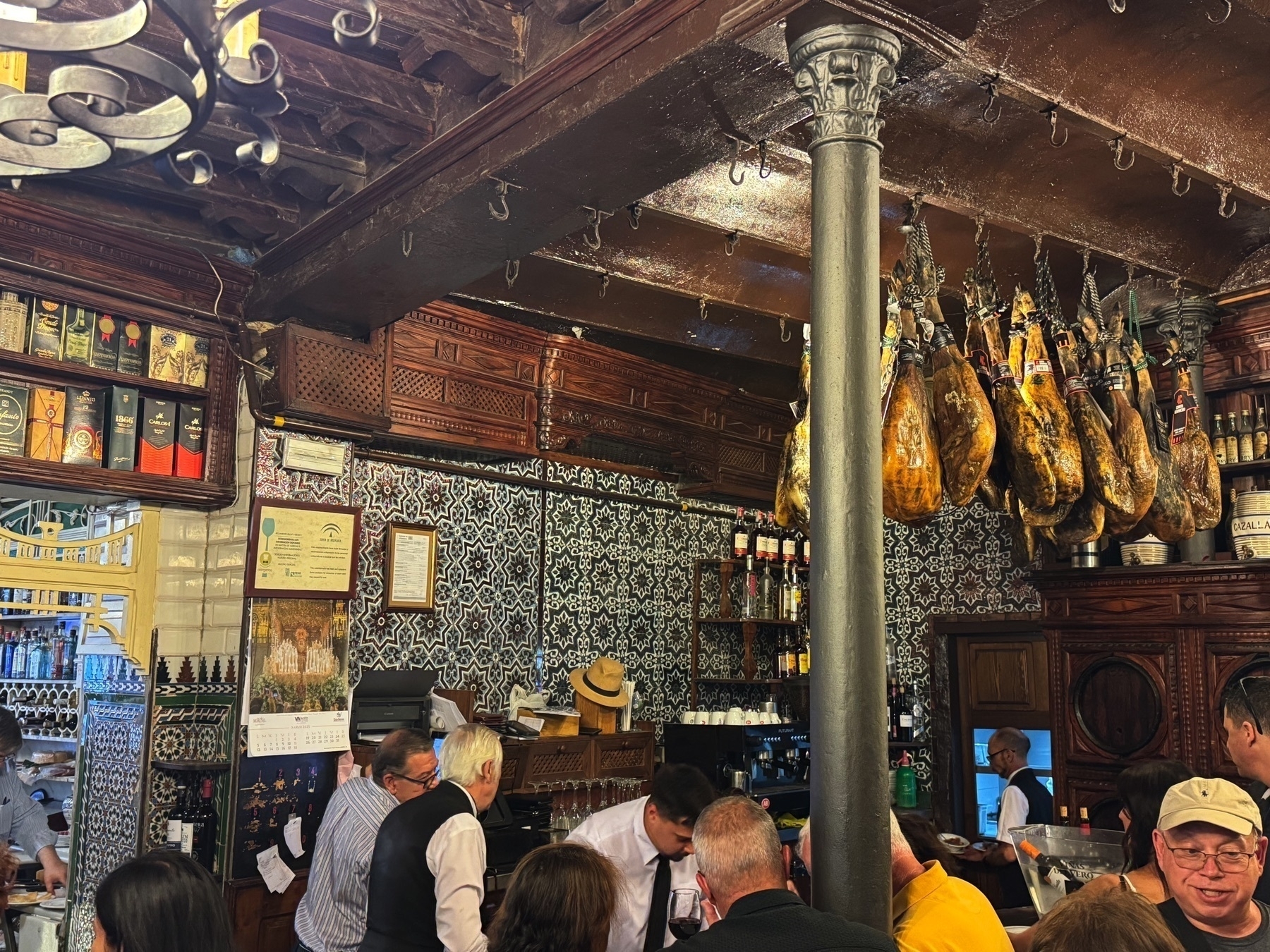 Bustling bar interior of El Rinconcillo with hanging jamón, ornate tiled walls, wood details, and patrons enjoying drinks, capturing the traditional Andalusian atmosphere.