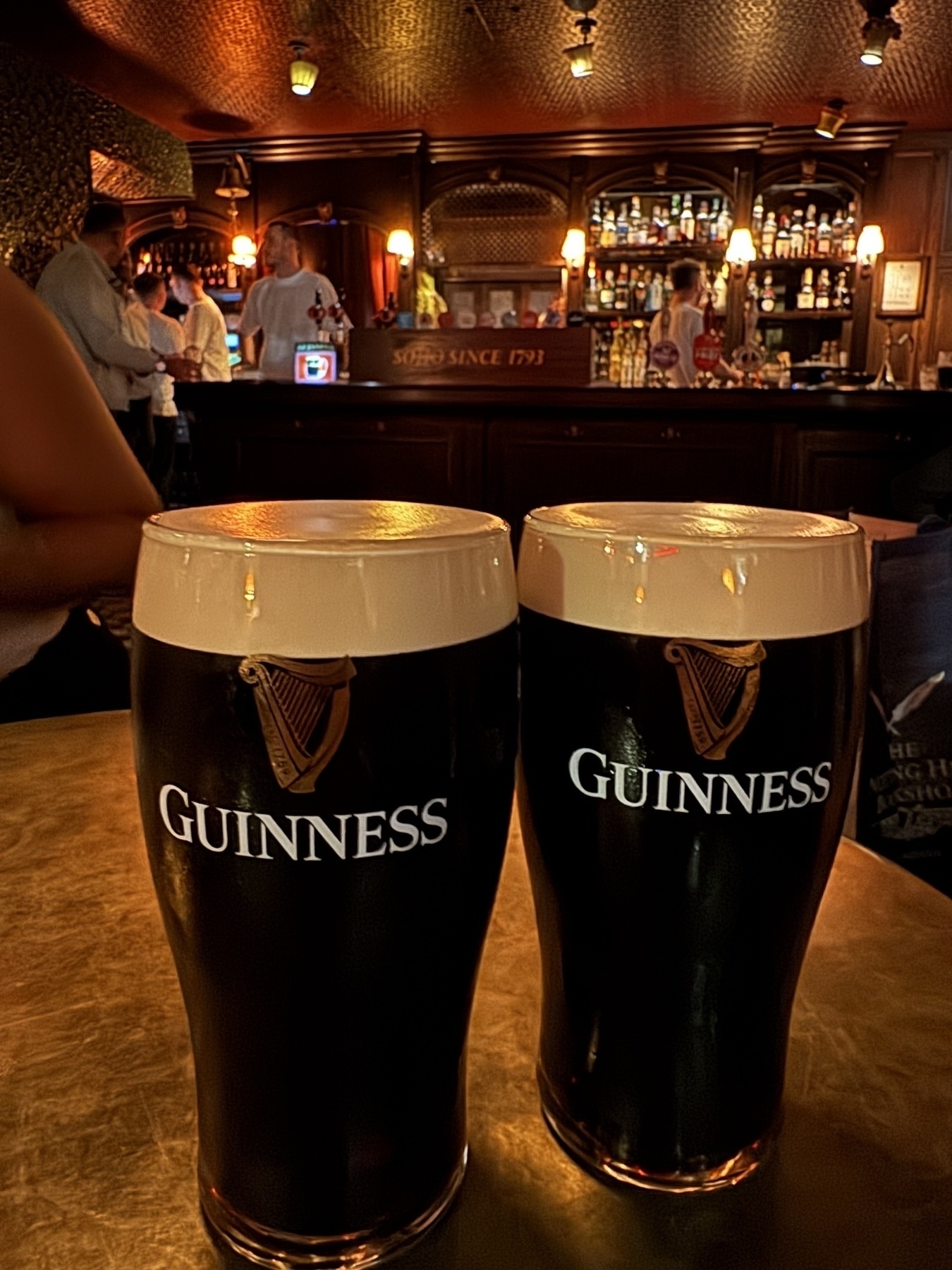 Two full Guinness pints with creamy heads sit on a gold-toned table in a warmly lit pub with a dark wood bar marked Soho Since 1793.
