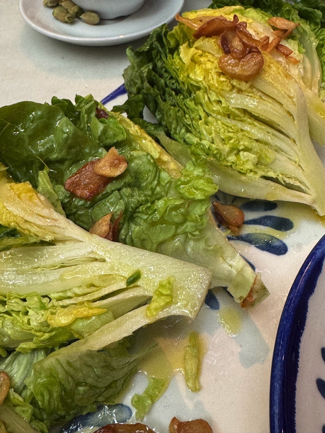 Wedges of fresh romaine lettuce lightly dressed with olive oil and topped with golden, crispy slices of garlic, arranged on a hand-painted ceramic plate with blue floral accents.