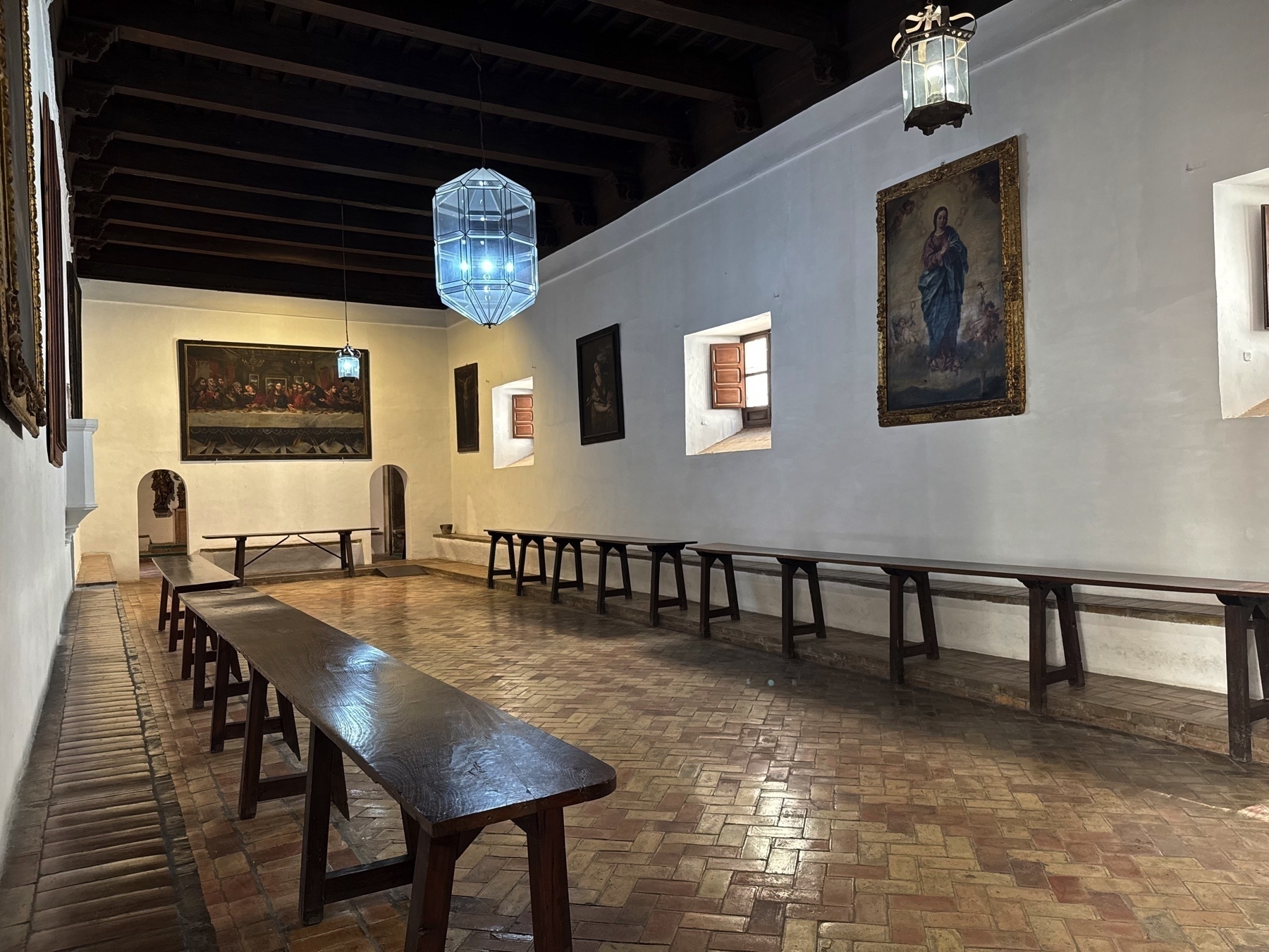 Simple monastic dining hall with long wooden tables and benches lining both sides of the room. Soft natural light enters through small square windows, highlighting the whitewashed walls and brick floor. Religious paintings, including a rendition of the Last Supper, adorn the walls beneath exposed wooden beams.