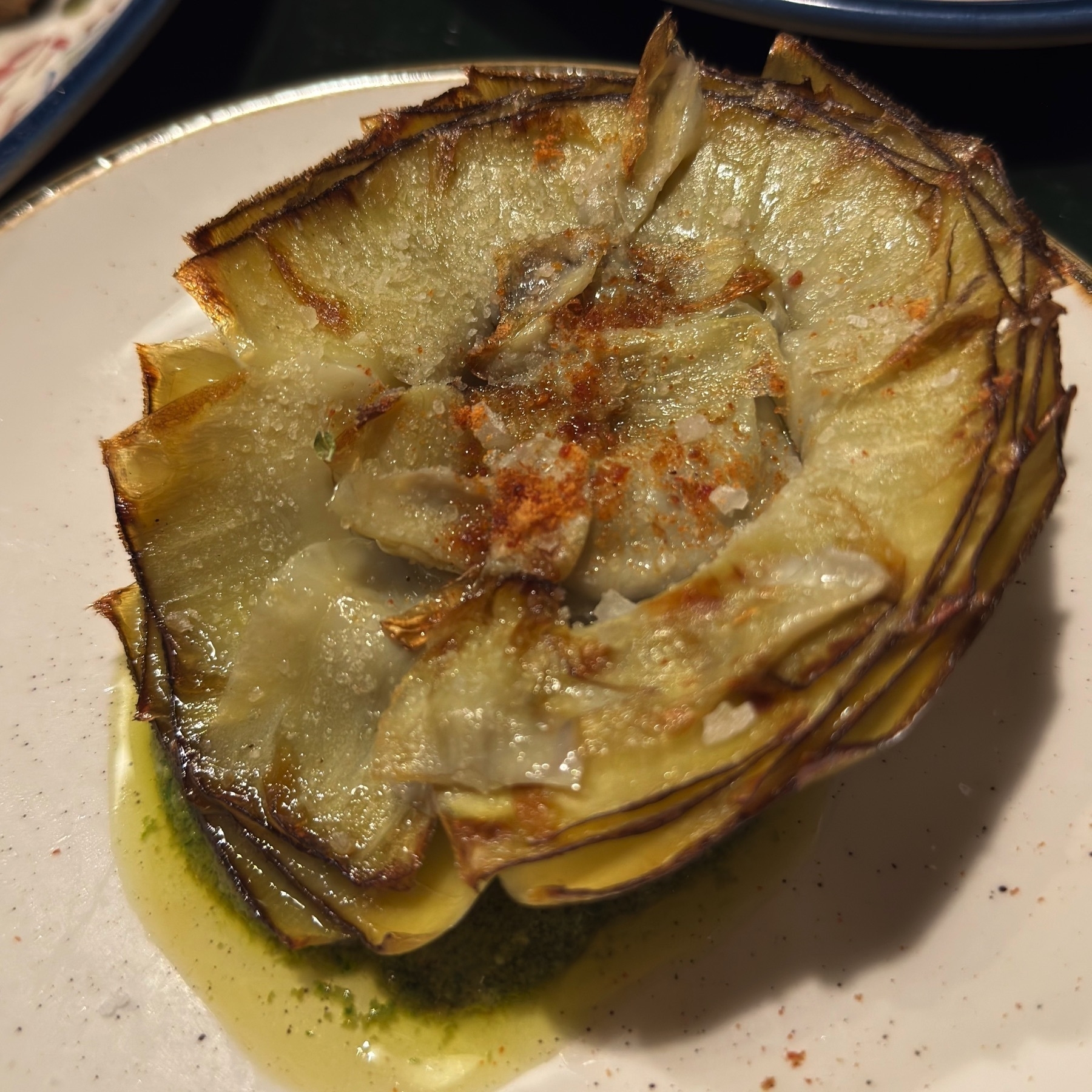 A beautifully presented fried artichoke, sliced thin and fanned out in a flower-like shape, with crisp, golden-brown edges and a tender, translucent interior. It glistens with olive oil and is sprinkled with coarse sea salt and paprika, creating an elegant balance of texture and seasoning.
