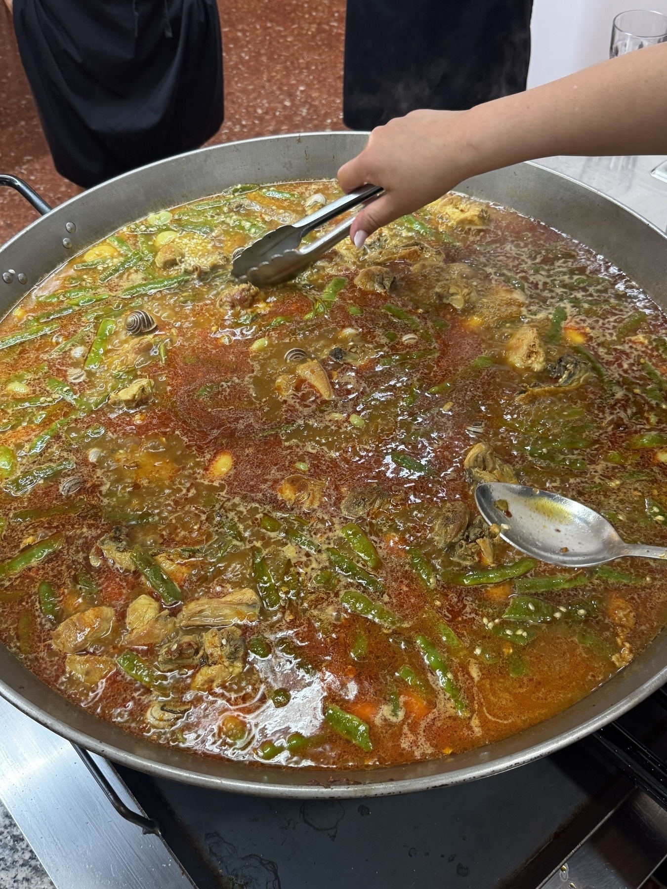 Paella cooking in broth with chicken, snails, and vegetables as someone uses tongs to adjust ingredients and a spoon to stir.