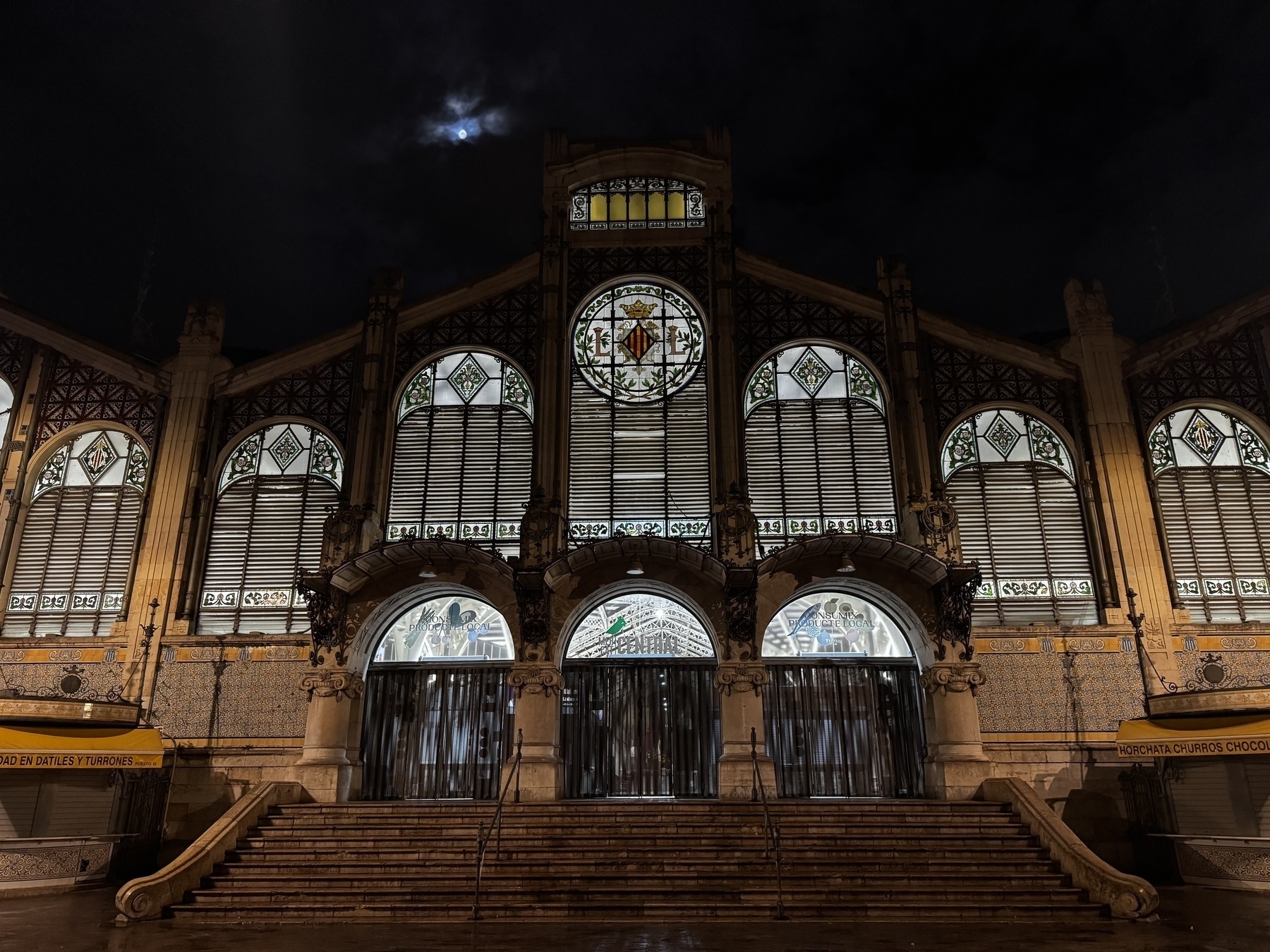 Nighttime view of the main entrance of Mercat Central de València, illuminated from within, with closed shutters, stained glass windows, and decorative tilework beneath a cloudy night sky and a partially visible moon.