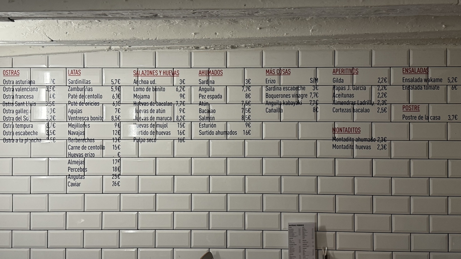Wall-mounted menu at Ostras Pedrín printed on white subway tiles, listing categories such as oysters, canned seafood, cured items, smoked items, and appetizers, with prices in euros.