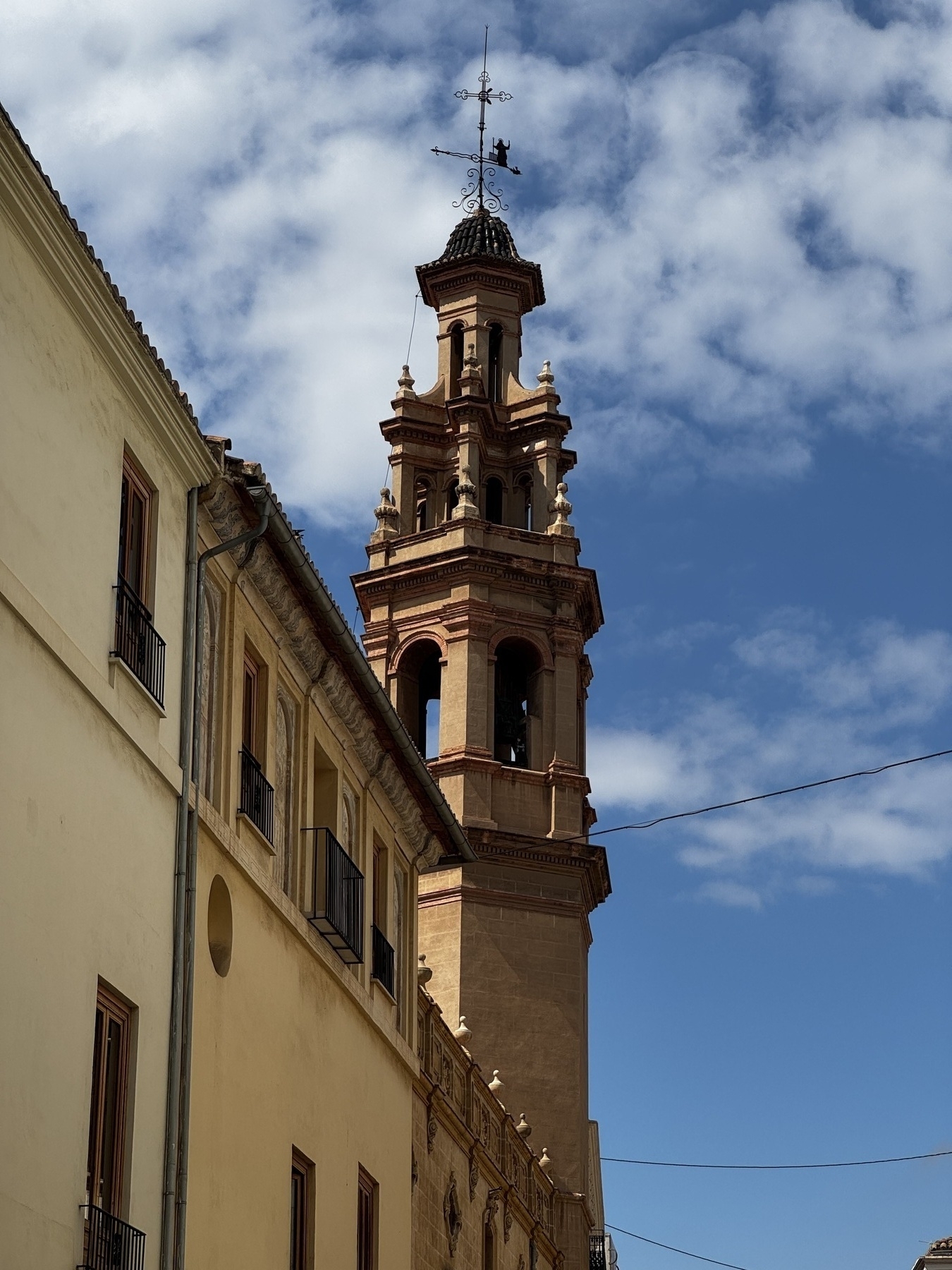 An elegant bell tower with terracotta details and a delicate wrought iron weather vane rises above an old monastery. The mix of warm stone and crisp light captures the charm of southern European architecture.