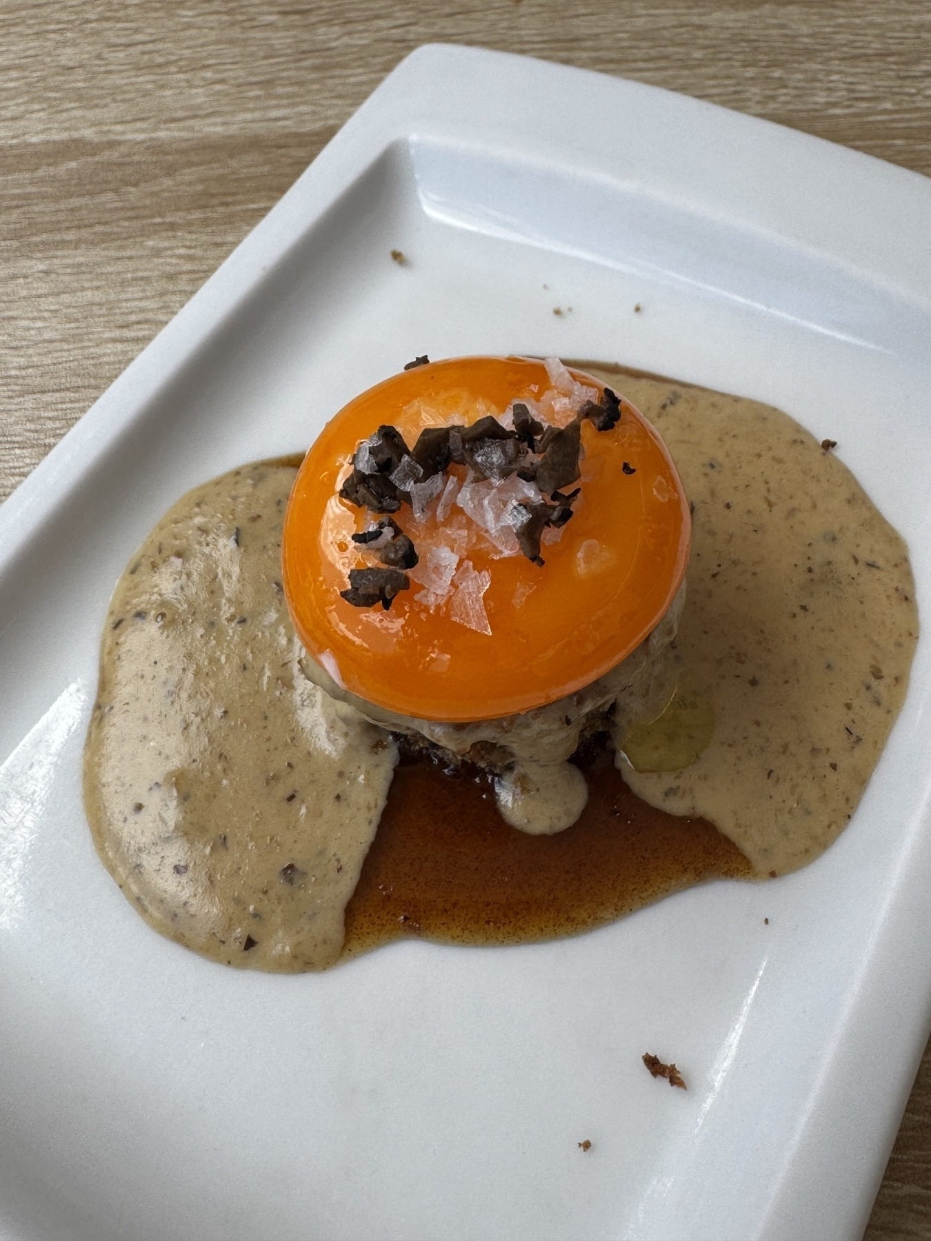 A silky egg yolk perched atop a moist mushroom sponge cake, with truffle-infused cream pooling around the base, and topped with crystals of salt and cacao nibs that add texture and umami depth.