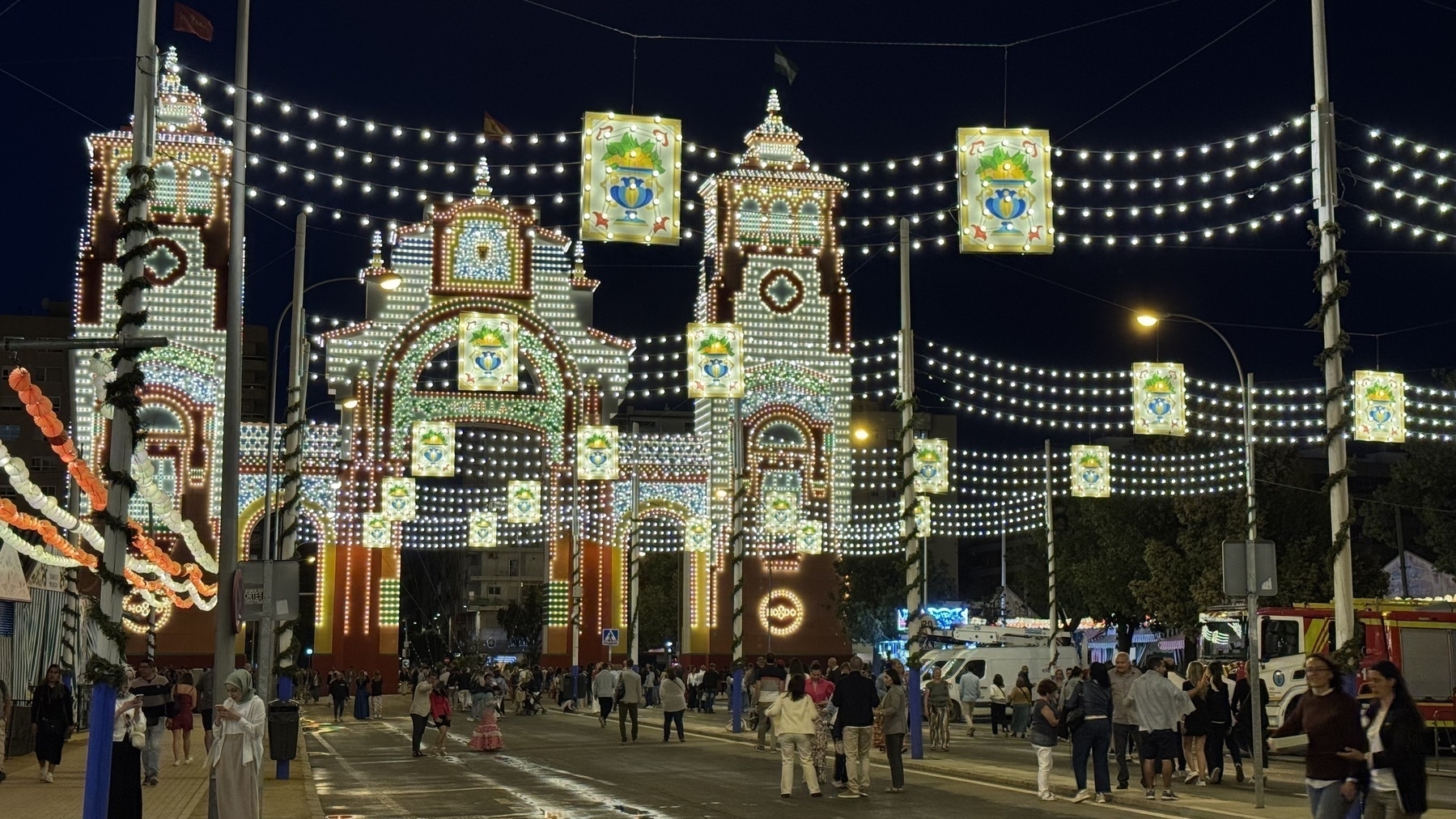 A large, illuminated archway adorned with intricate light patterns and framed by strings of lanterns overhead. People walk beneath the glowing entrance, enhancing the sense of arrival and celebration.