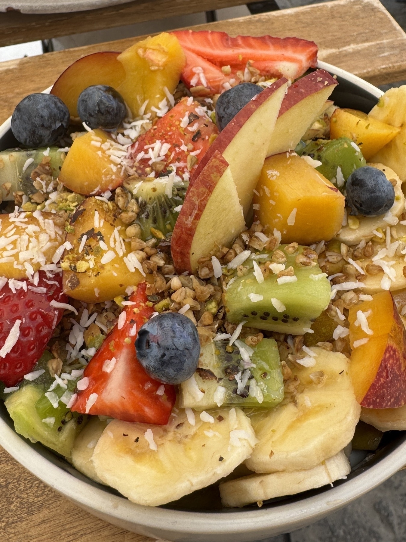 A bright and colorful fruit bowl from Parcería Café filled with sliced bananas, strawberries, apple wedges, kiwi chunks, pineapple, and fresh blueberries. Topped with granola and shredded coconut, the bowl offers a mix of textures and vibrant colors that make it look nourishing, flavorful, and satisfying for a wholesome breakfast.