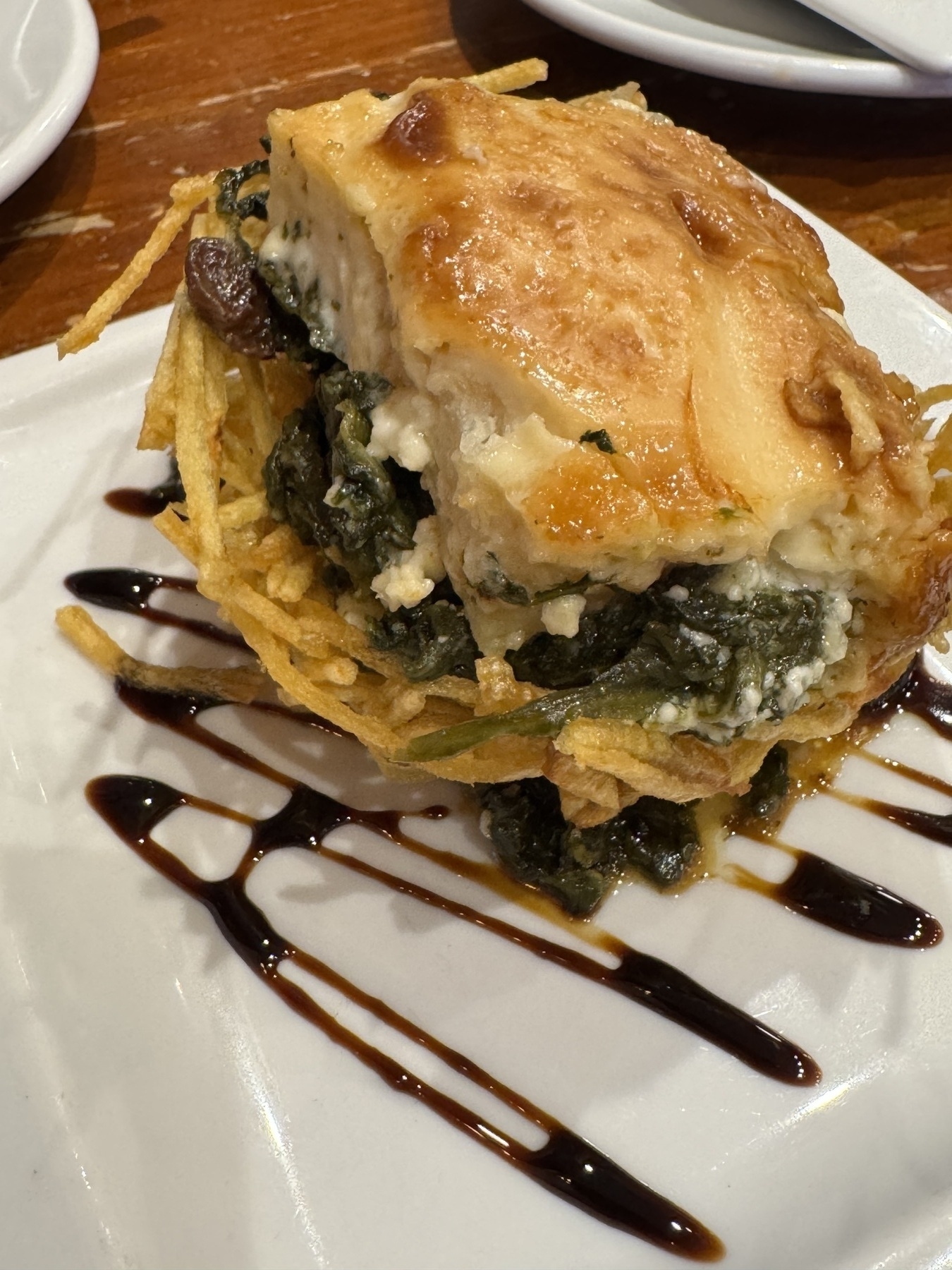 A carefully layered timbale identified as Nido de Bacalao y Espinacas. The base is formed from thin, crispy potato threads, supporting layers of spinach and flaky cod, all topped with a golden crust of baked béchamel or cheese. A balsamic reduction surrounds the dish, adding a glossy, elegant touch.