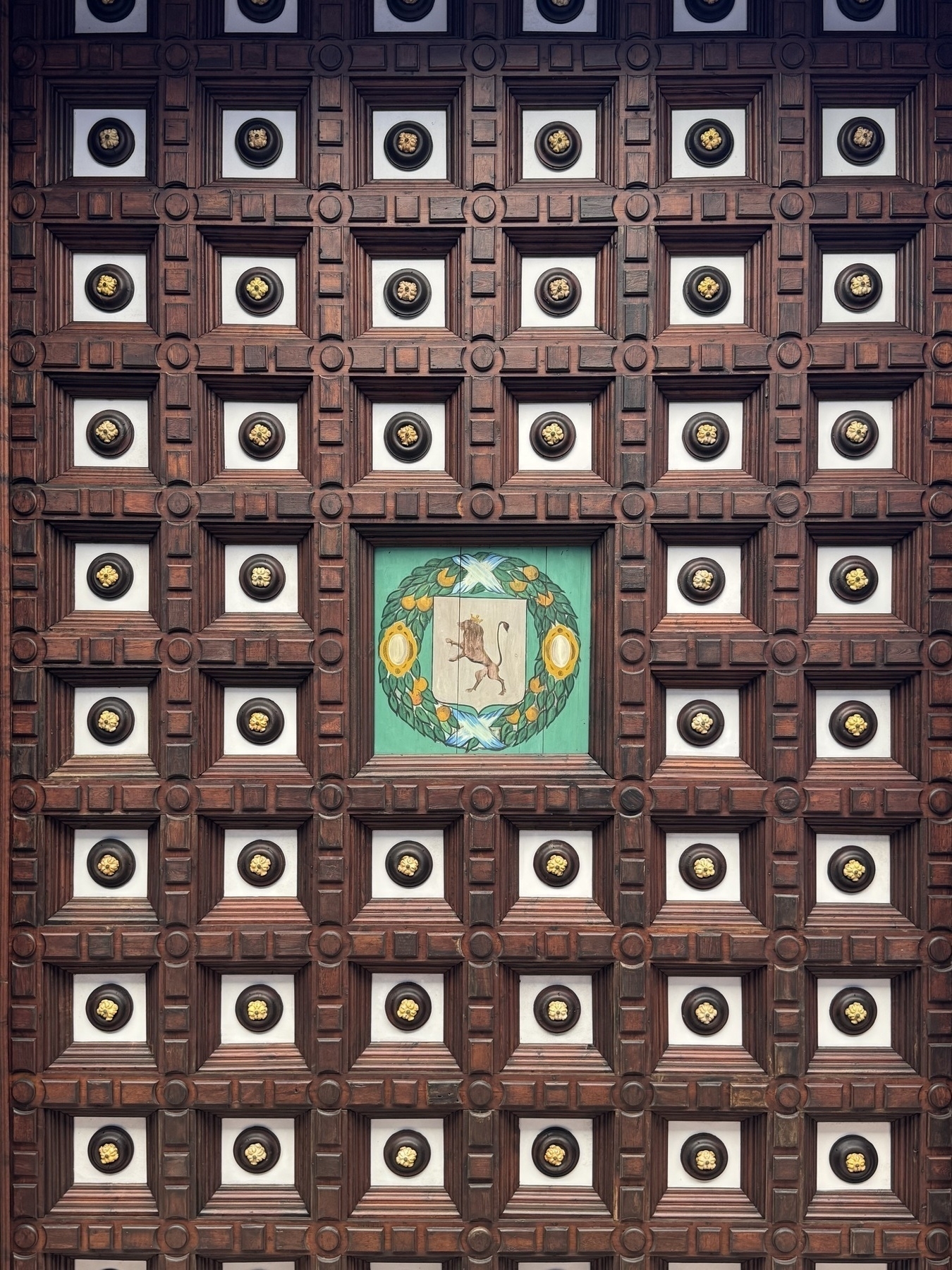 An intricately carved wooden ceiling composed of square panels, each containing a gold emblem on a white background. The central panel is painted green with a crest showing a deer surrounded by a laurel wreath, encircled by fruit, ribbon, and stylized leaves.