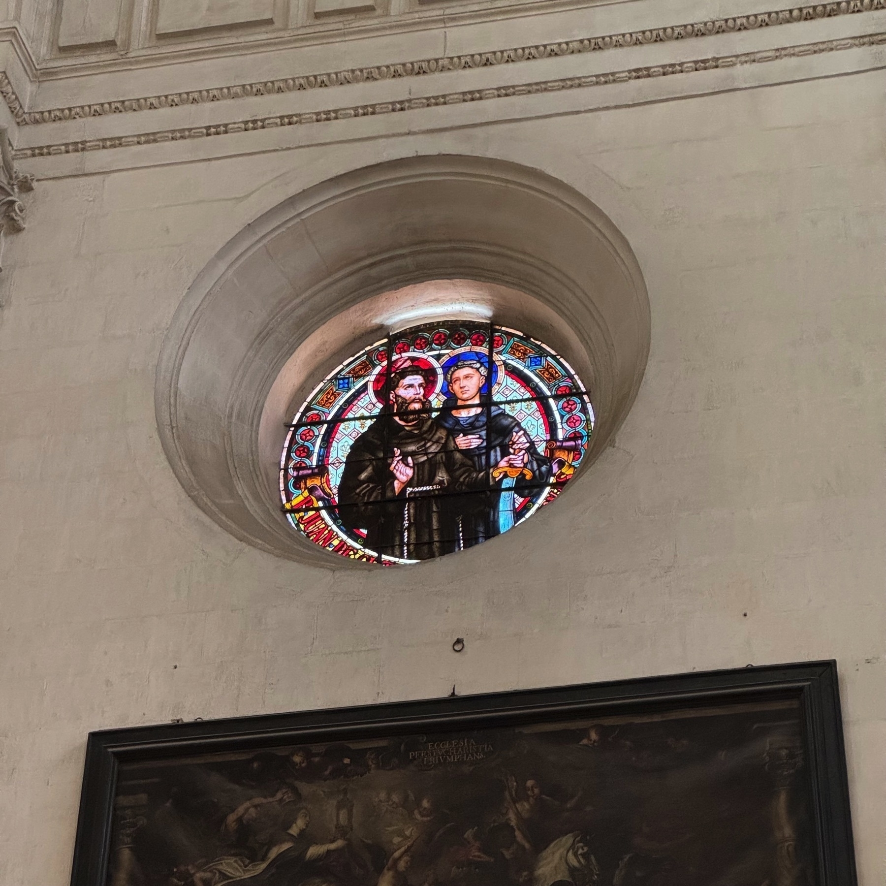 Circular stained-glass window featuring two male religious figures, one older and bearded in brown robes, the other younger in blue. The window is inset into a plain white wall with minimal decoration, allowing the vibrant colors and expressive detail of the glasswork to stand out dramatically against the surrounding simplicity.