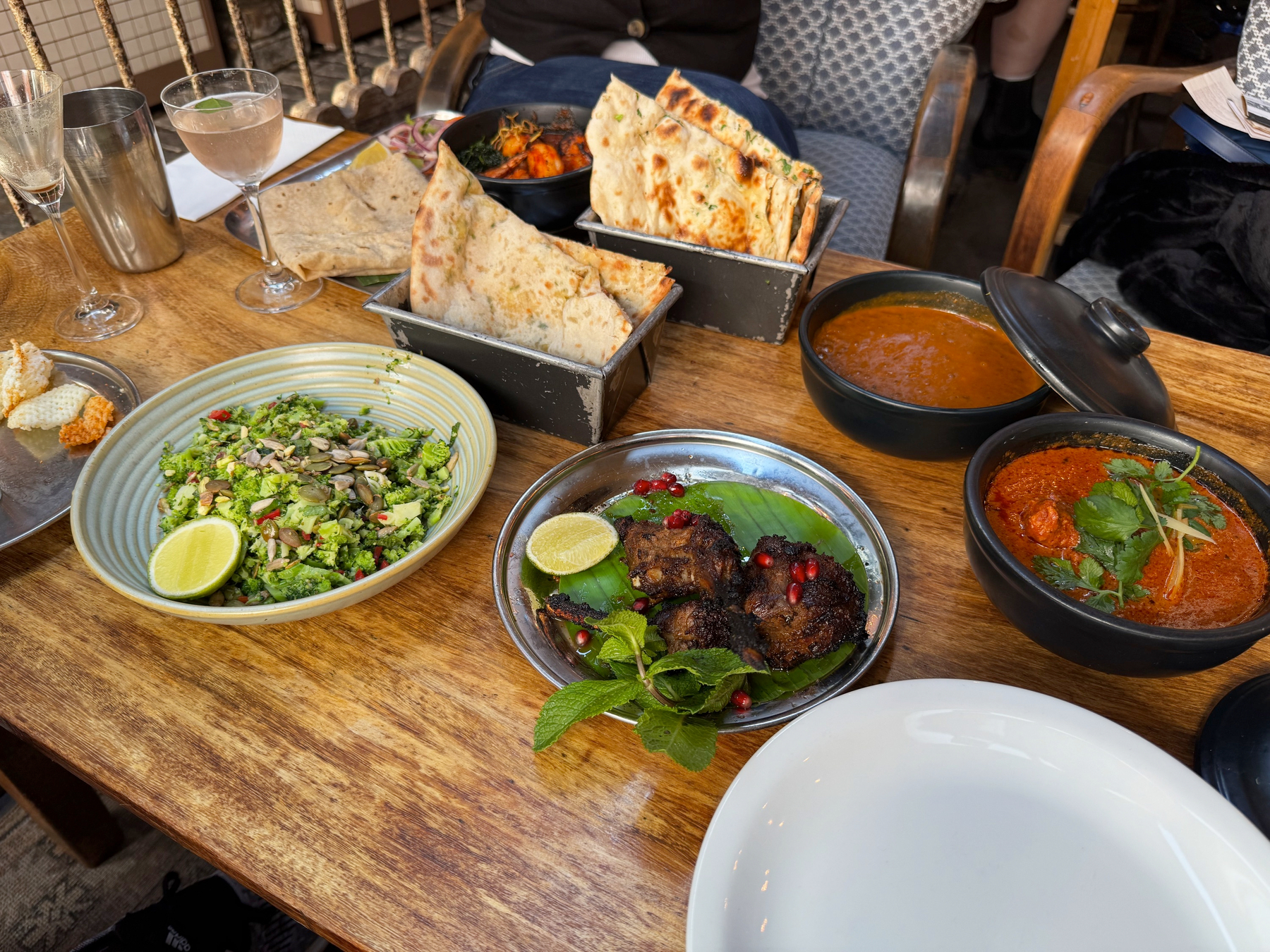 A generous spread of Indian dishes fills a wooden table, with blistered naan stacked in metal tins, bowls of rich red curry glistening with oil, a bright green chopped salad scattered with seeds, and charred lamb chops resting on a banana leaf with lime and mint. The mix of textures, colors, and shared plates creates a warm, abundant, and inviting dining scene.
