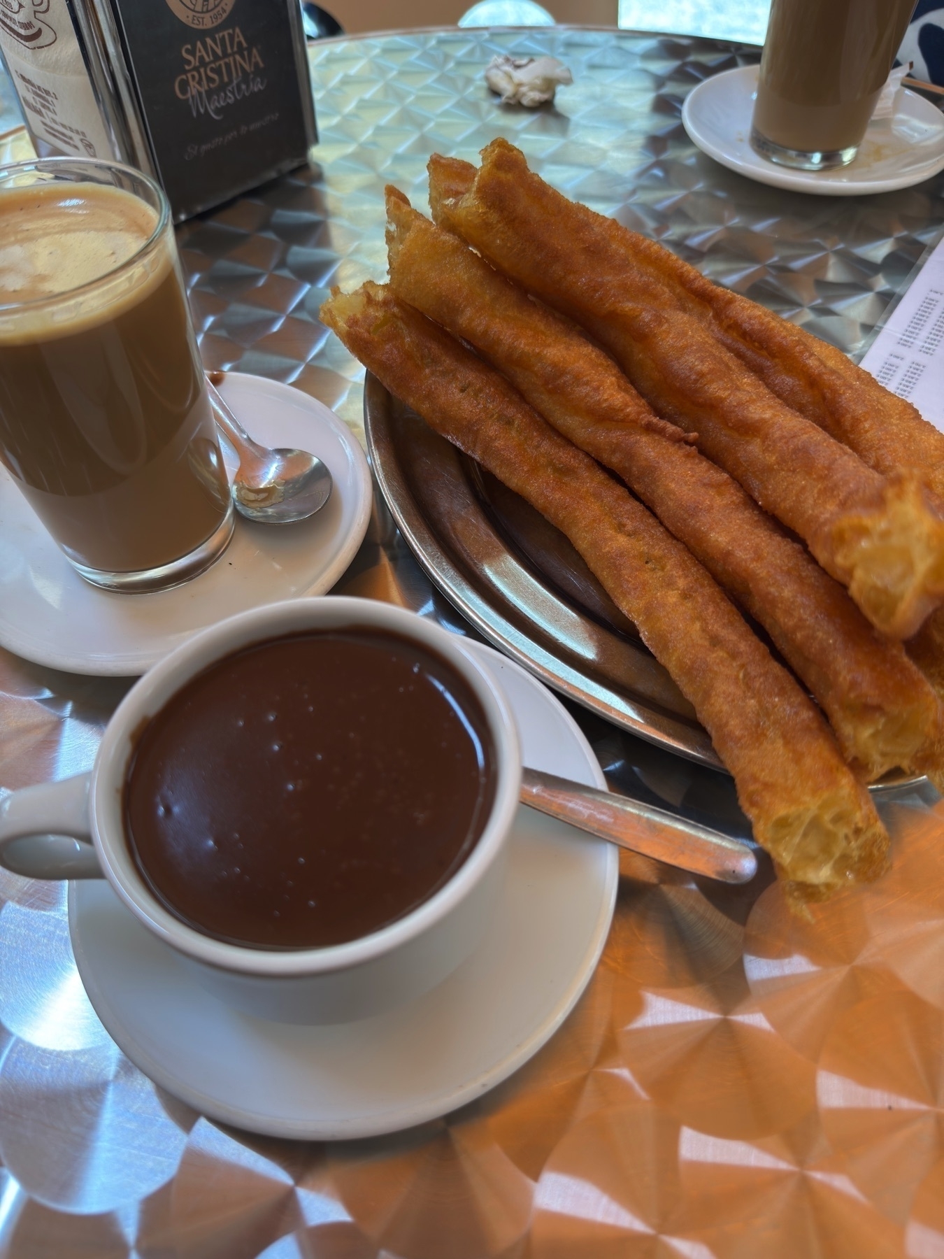 A classic Spanish breakfast of crisp, golden churros on a metal plate, served with a thick cup of hot chocolate and a glass of café con leche. The churros look light and airy, ideal for dipping in the rich chocolate.