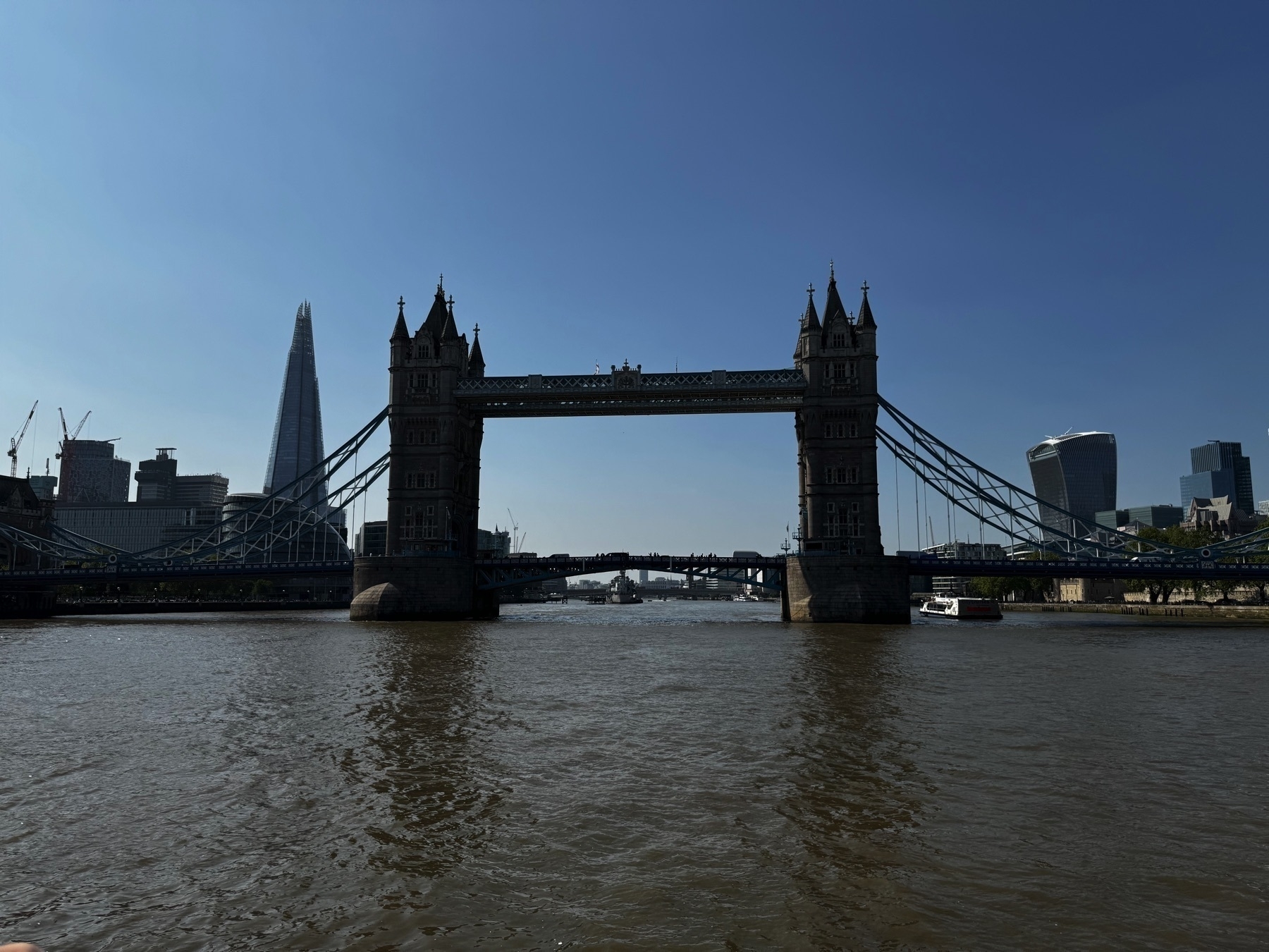 A wide view of Tower Bridge spanning the River Thames on a clear day, with the sharp silhouette of the Shard on the left and the curved Walkie-Talkie building on the right. The river stretches calmly into the distance.