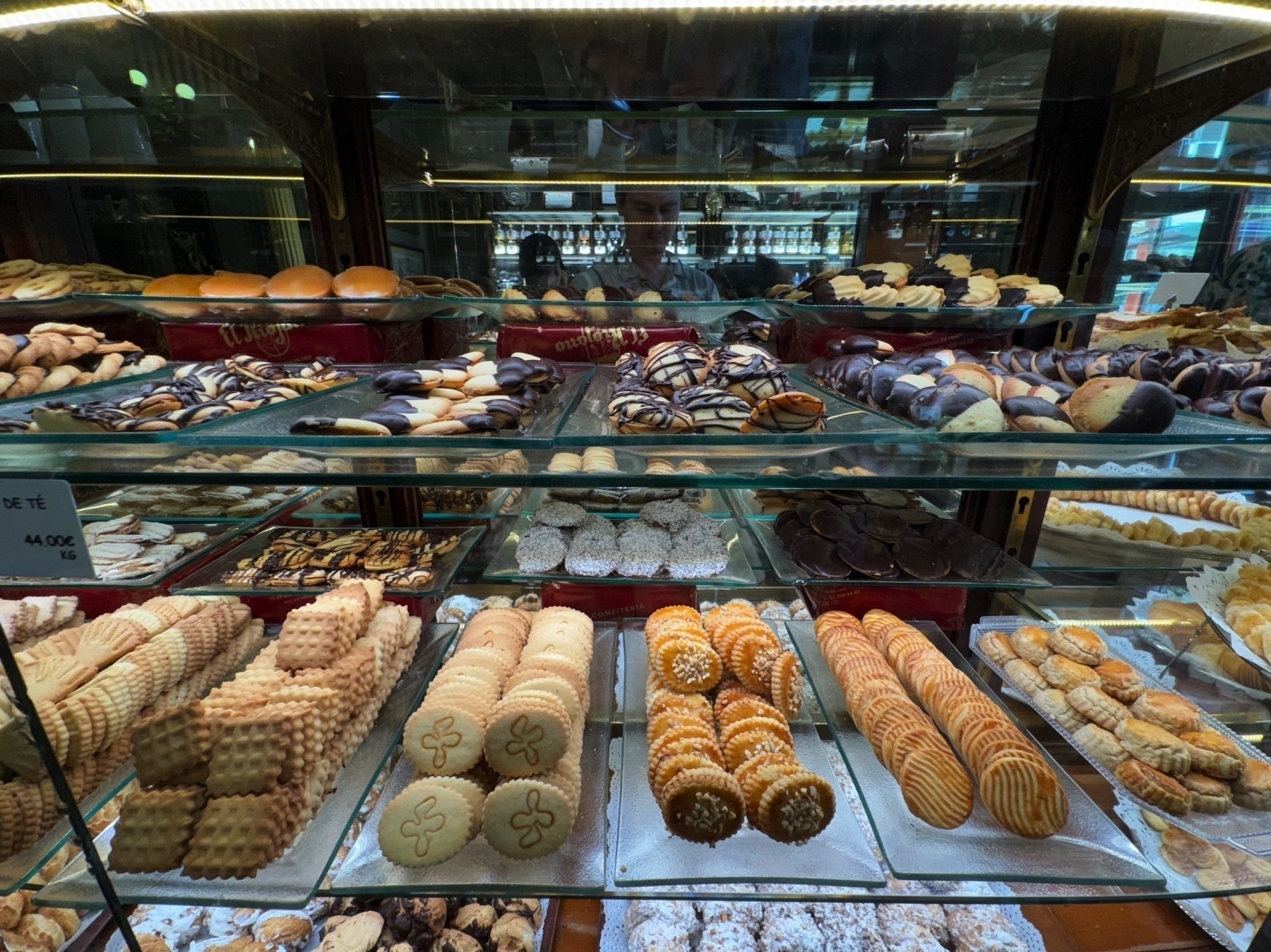 Rows of assorted cookies and pastries fill a glass display, organized by type and color in visually pleasing symmetry. The front row features geometric butter cookies, golden spirals, and nut-topped rounds, while the background highlights chocolate-dipped biscuits and striped pastries. The reflection of customers adds a candid charm to the elegant setting, capturing the moment of choice at a classic pastry counter.