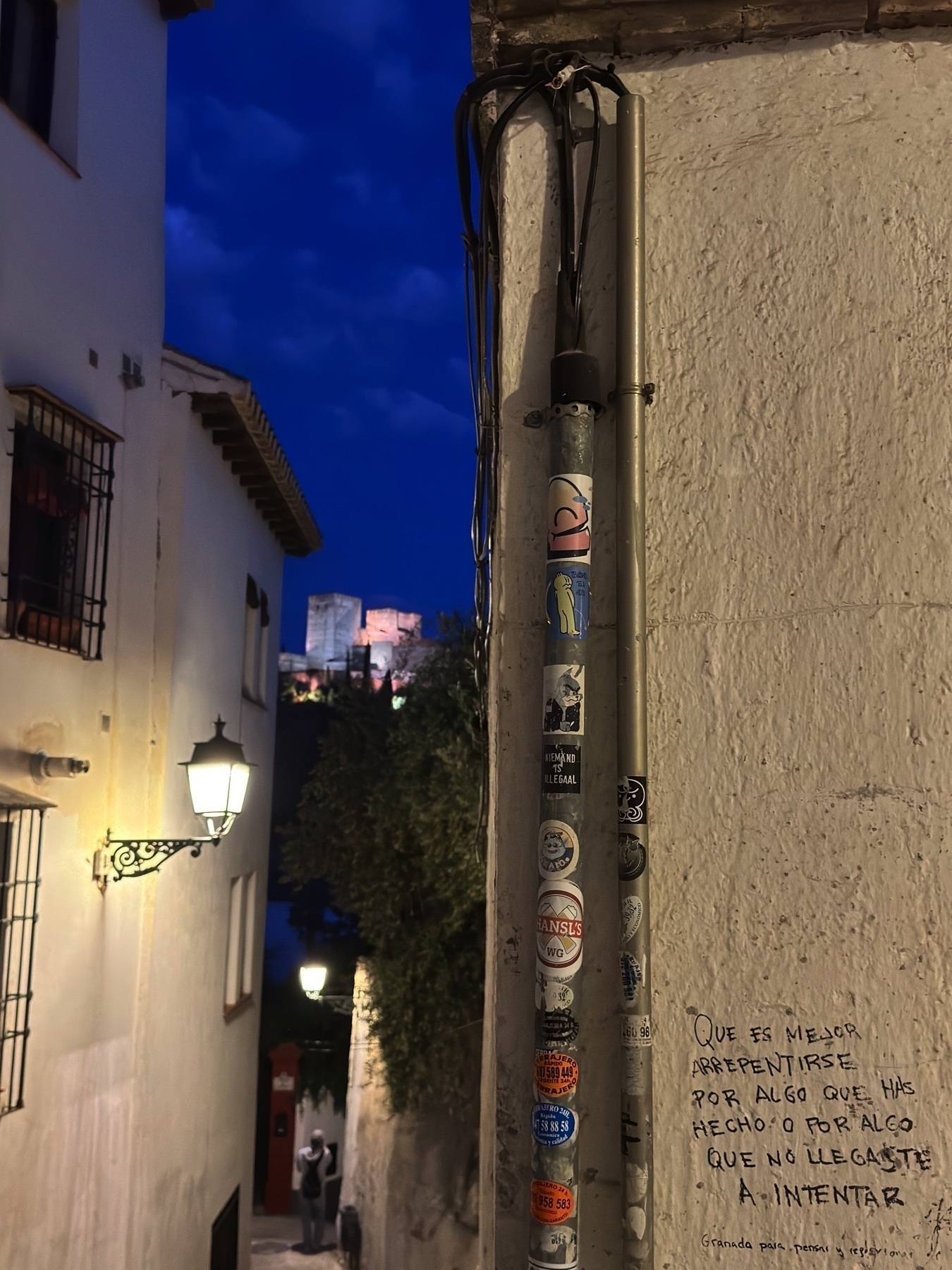 Narrow alleyway leading downhill with the Alhambra visible in the distance, softly lit against the night sky. A wall-mounted light casts a glow on the whitewashed buildings, and a utility pole covered in stickers is in sharp focus beside a wall with handwritten graffiti in Spanish.