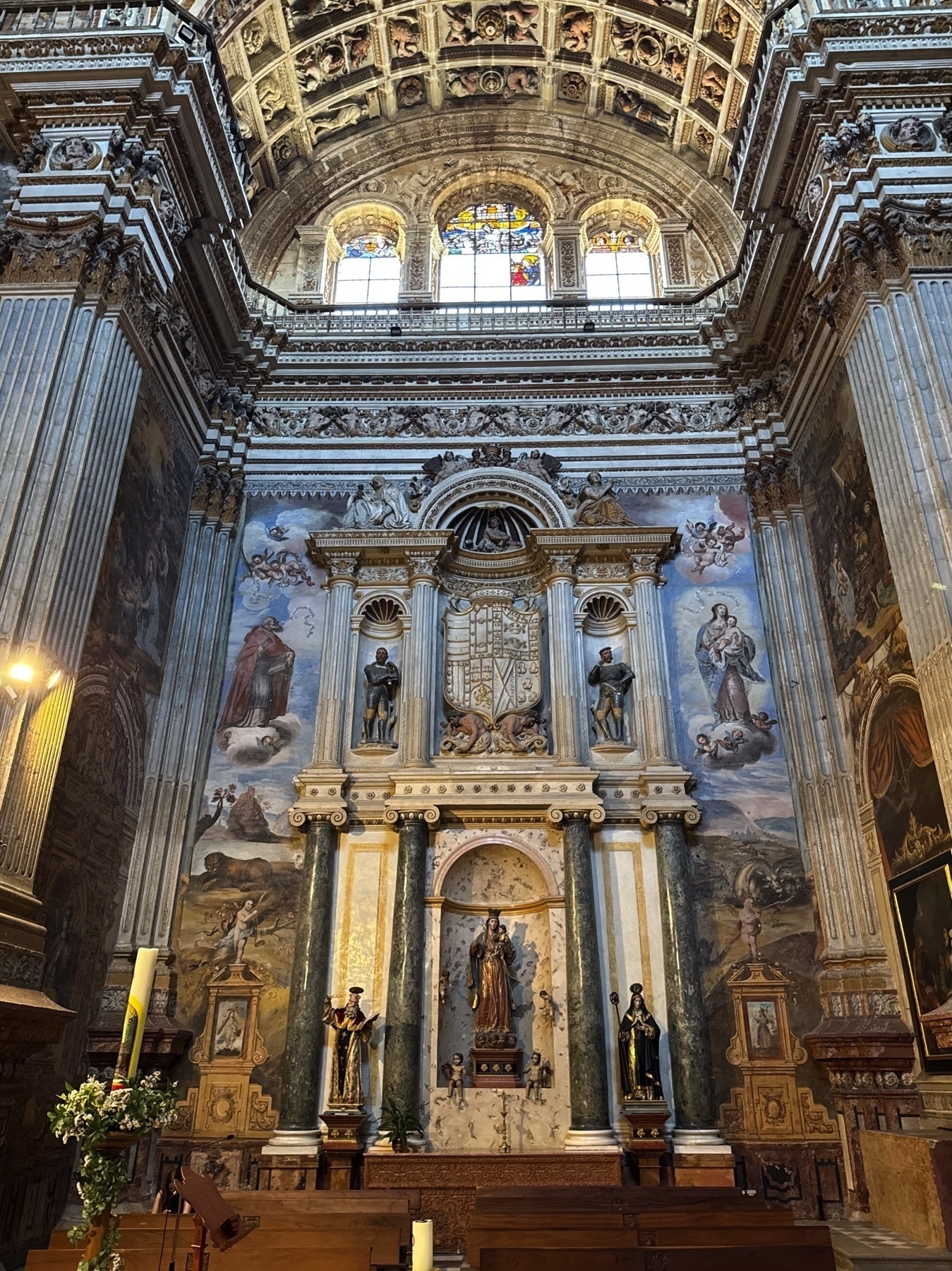 Altarpiece framed by soaring columns and detailed frescoes, featuring statues of saints and biblical figures set against a painted backdrop. Above, three arched stained glass windows illuminate the richly decorated apse, bringing light and color into the sacred space.