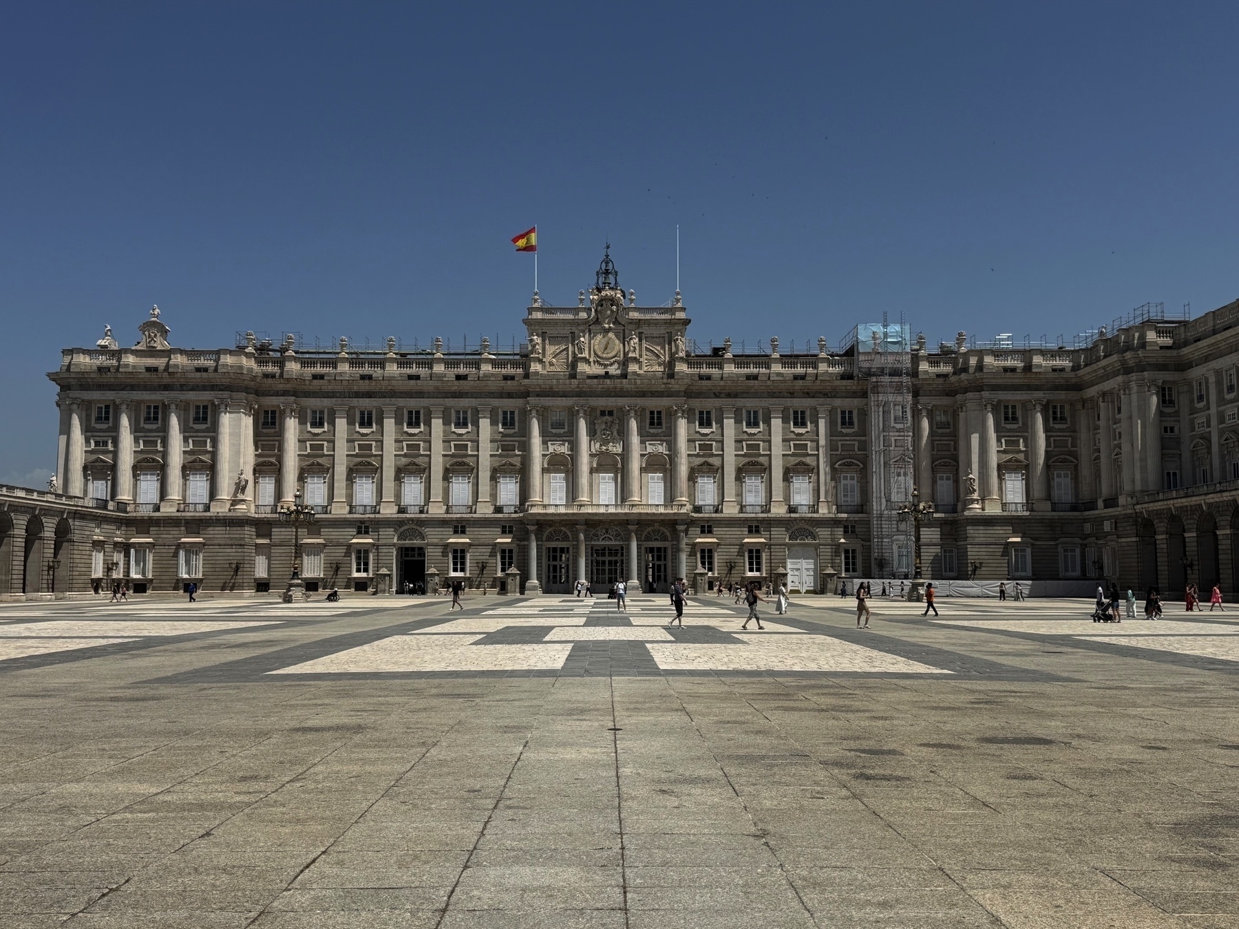 Wide-angle view of the Royal Palace of Madrid, with its grand neoclassical façade featuring rows of tall columns, numerous windows, and ornate sculptures. The Spanish flag flies prominently from the roof. The expansive forecourt in front of the building is paved with geometric stone patterns, and scattered visitors walk or stand, giving a sense of scale to the immense structure. The sky is clear and blue, enhancing the elegance and formality of the architecture.