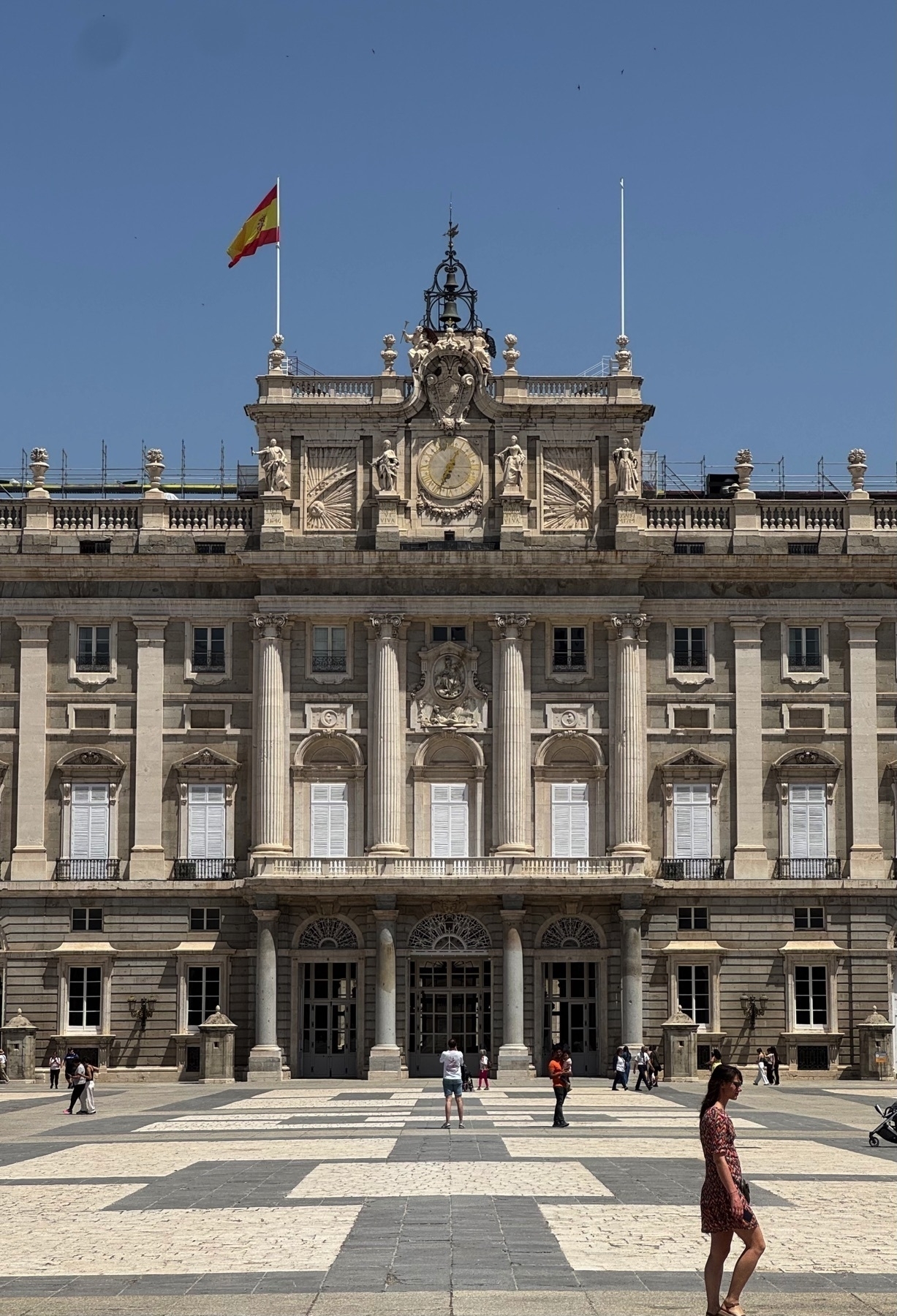 Closer view of the central section of the Royal Palace of Madrid, highlighting the symmetrical stonework, decorative columns, and arched doorways. A large clock is set into the ornate pediment above the entrance, flanked by classical statues. The Spanish flag waves atop the building, and several people move across the stone courtyard in the foreground, some pausing to admire the architecture.