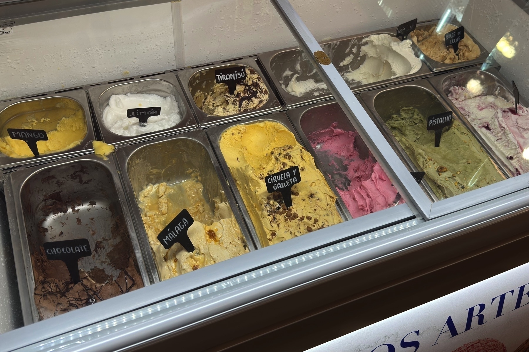 A freezer case of artisanal ice cream showcases over a dozen flavors in stainless steel tubs, each labeled with handwritten signs. Vibrant colors—from mango yellow and pistachio green to deep chocolate brown and bright raspberry pink—suggest creamy, rich textures and diverse taste profiles. The inviting presentation emphasizes freshness and variety, ideal for sampling on a warm day.