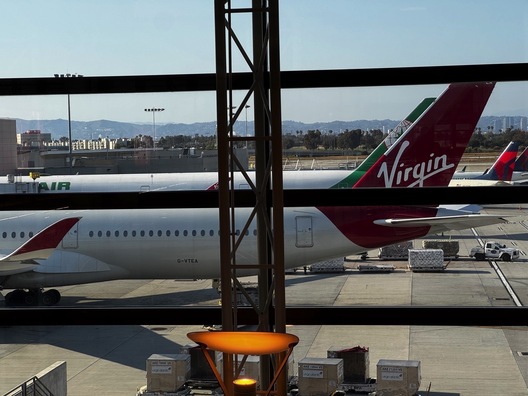 Virgin Atlantic aircraft parked at the gate, seen through large terminal windows. The plane's red and green tail fin with the Virgin logo is prominently visible, with other airline tails and airport ground equipment in view.