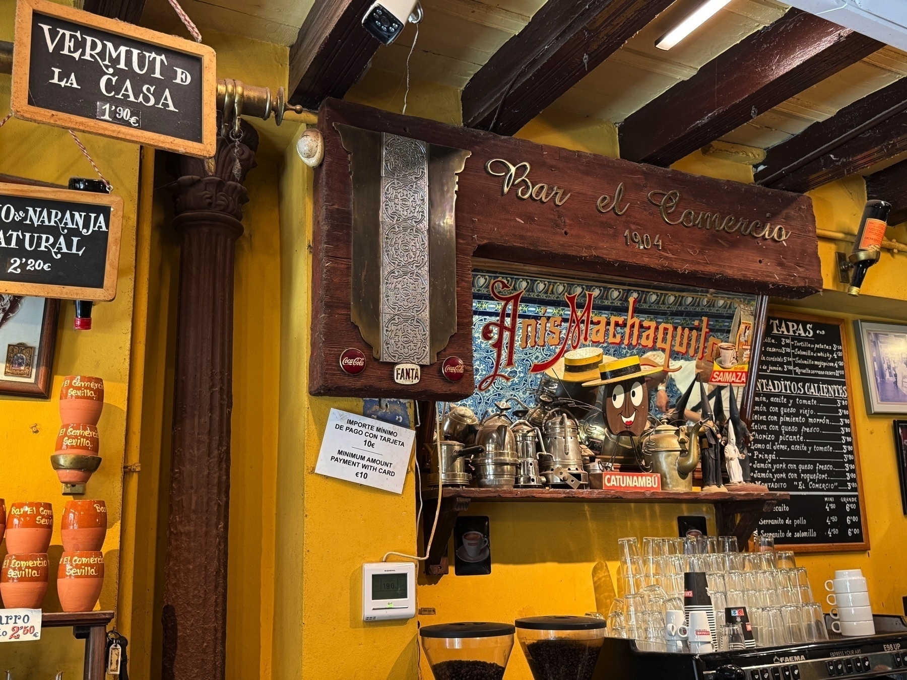 The cozy interior of Bar El Comercio features a vintage espresso setup with shiny metal coffee pots, a mirrored sign advertising Anís Machaquito, and blackboards listing house vermouth, natural orange juice, and tapas. The space is painted in bright yellow with wooden beams and rustic decor that evoke a classic Andalusian bar atmosphere.