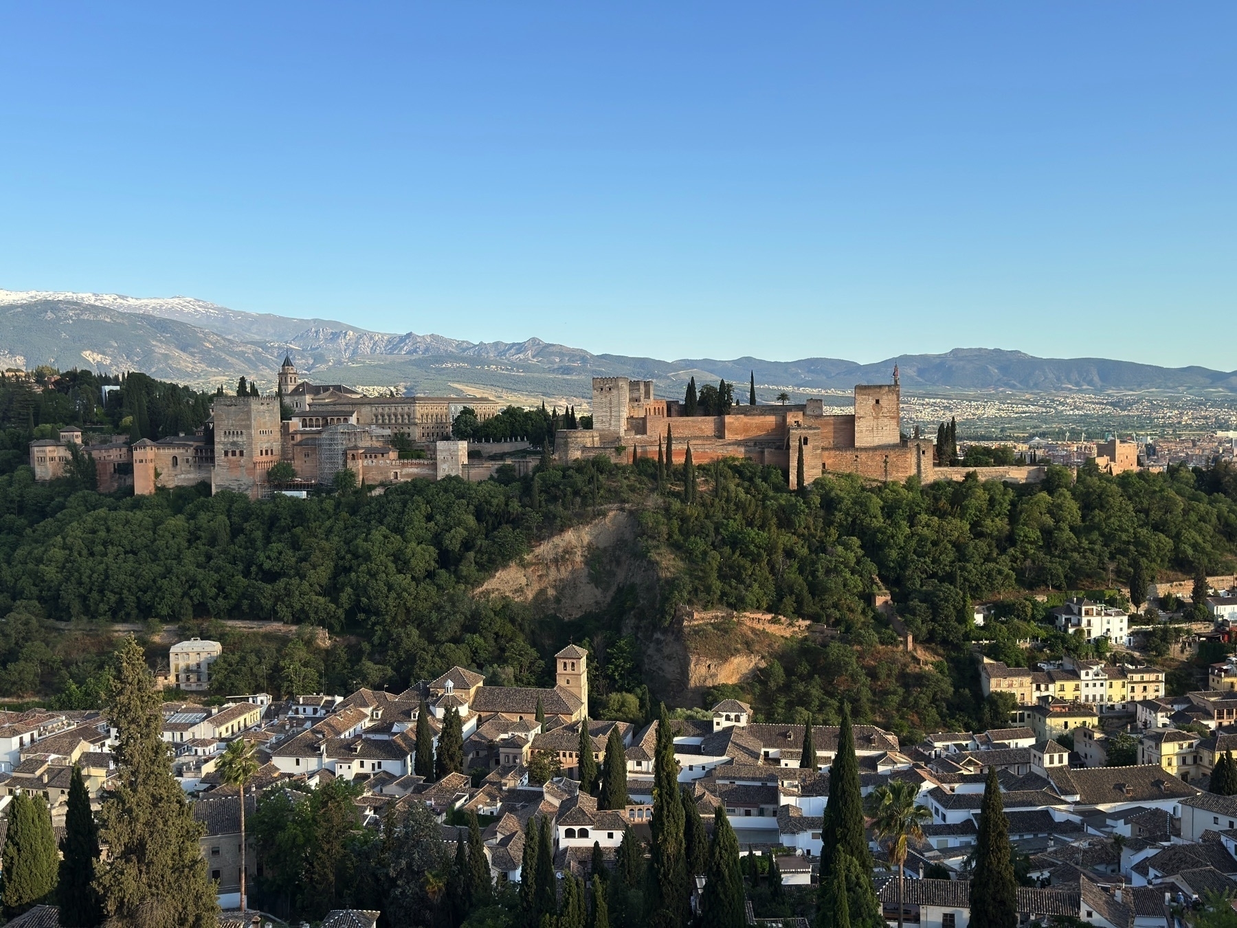 Wide shot of the Alhambra complex on a wooded hill, with its full array of palaces, towers, and defensive walls clearly visible. The foreground includes white buildings and cypress trees of the Albaicín neighborhood, with the Sierra Nevada mountains in the distance.