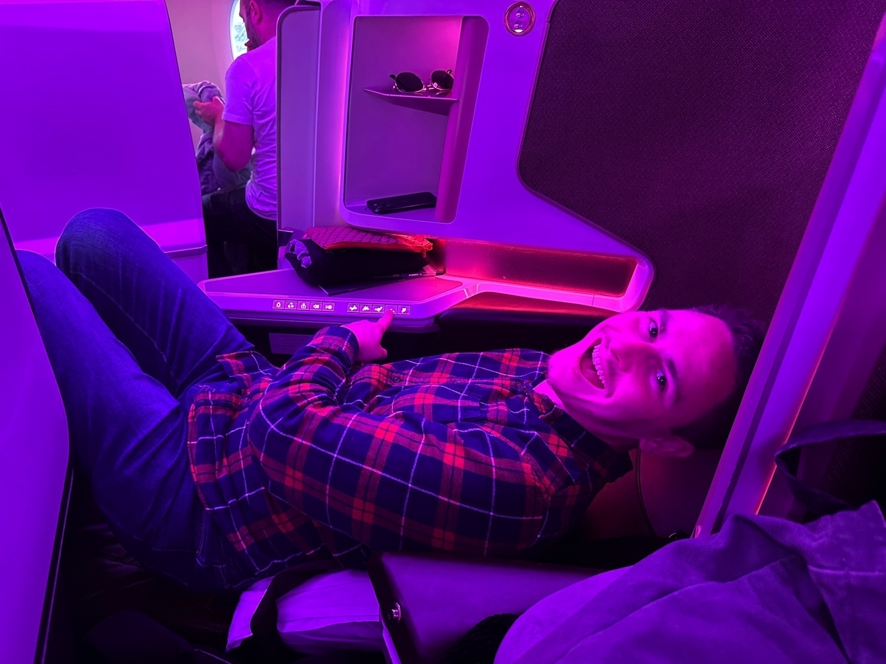 Man reclining and smiling in a lie-flat seat on a Virgin Atlantic flight, bathed in ambient purple lighting. The seat's console, entertainment screen, and personal storage compartments are visible around him.