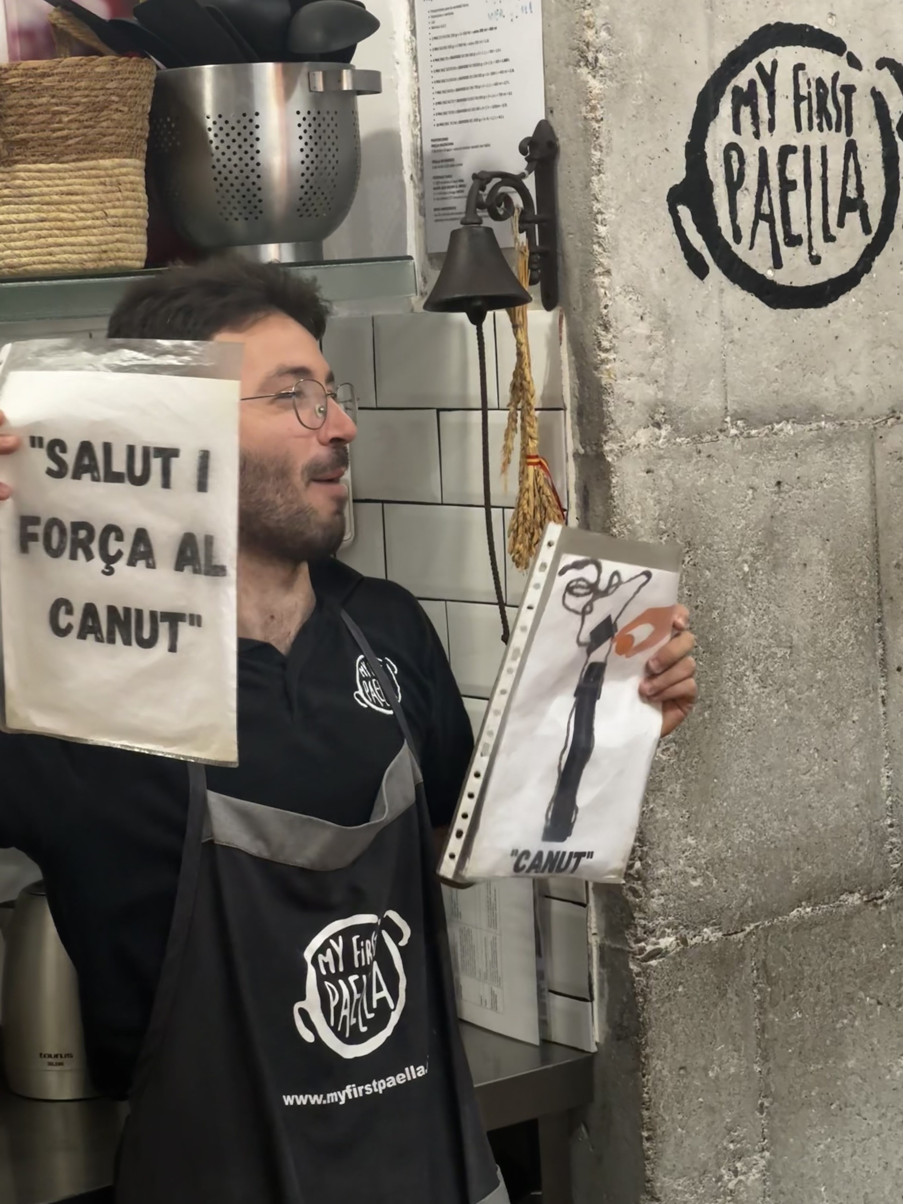 An instructor holding up a Catalan phrase sign reading “Salut i força al canut” and a humorous drawing of kitchen tongs labeled “Canut.”