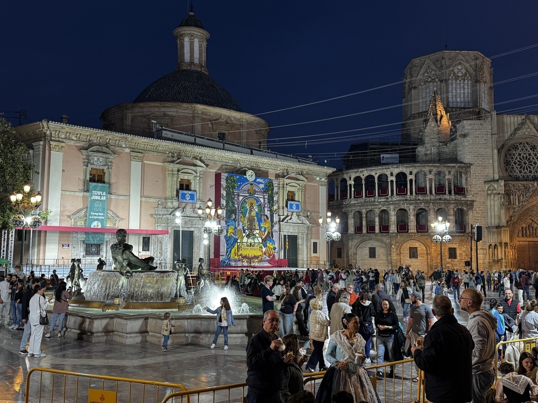 A vibrant plaza bustles with people at night, gathered around a fountain and a colorful floral display on a church façade. The festive atmosphere and dramatic lighting bring the square to life with energy and tradition.