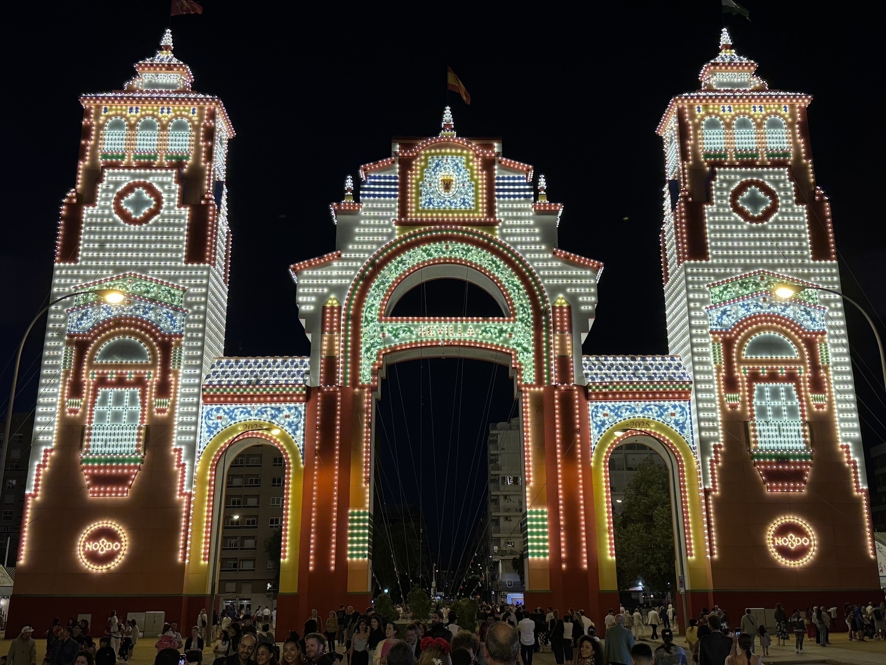 A close-up view of the main entrance arch lit up in red, green, white, and blue patterns with three towers. The crowd beneath the grand structure emphasizes its scale and iconic design.