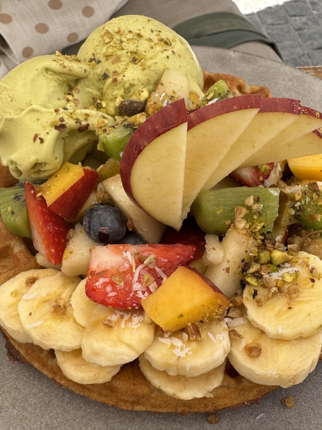 Close-up of a dessert plate from Parcería Café featuring a waffle base loaded with sliced banana, apple, kiwi, and strawberries, topped with scoops of pistachio-green ice cream. A sprinkling of granola and shredded coconut adds crunch and contrast, making the dish look indulgent yet fresh and perfect for a sweet brunch.