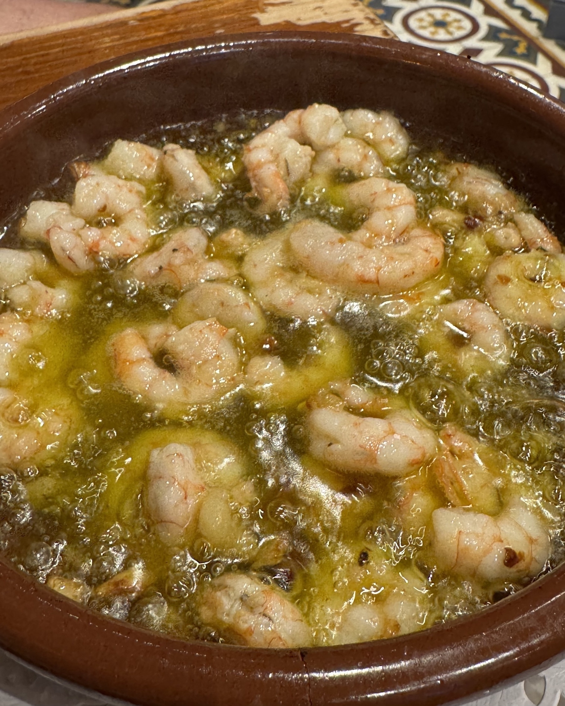 A rustic clay dish filled with bubbling gambas al ajillo—shrimp sizzling in hot garlic-infused olive oil. The shrimp are plump and slightly curled, surrounded by visible slivers of garlic and chili flakes. The intense bubbling emphasizes freshness and immediate serving.