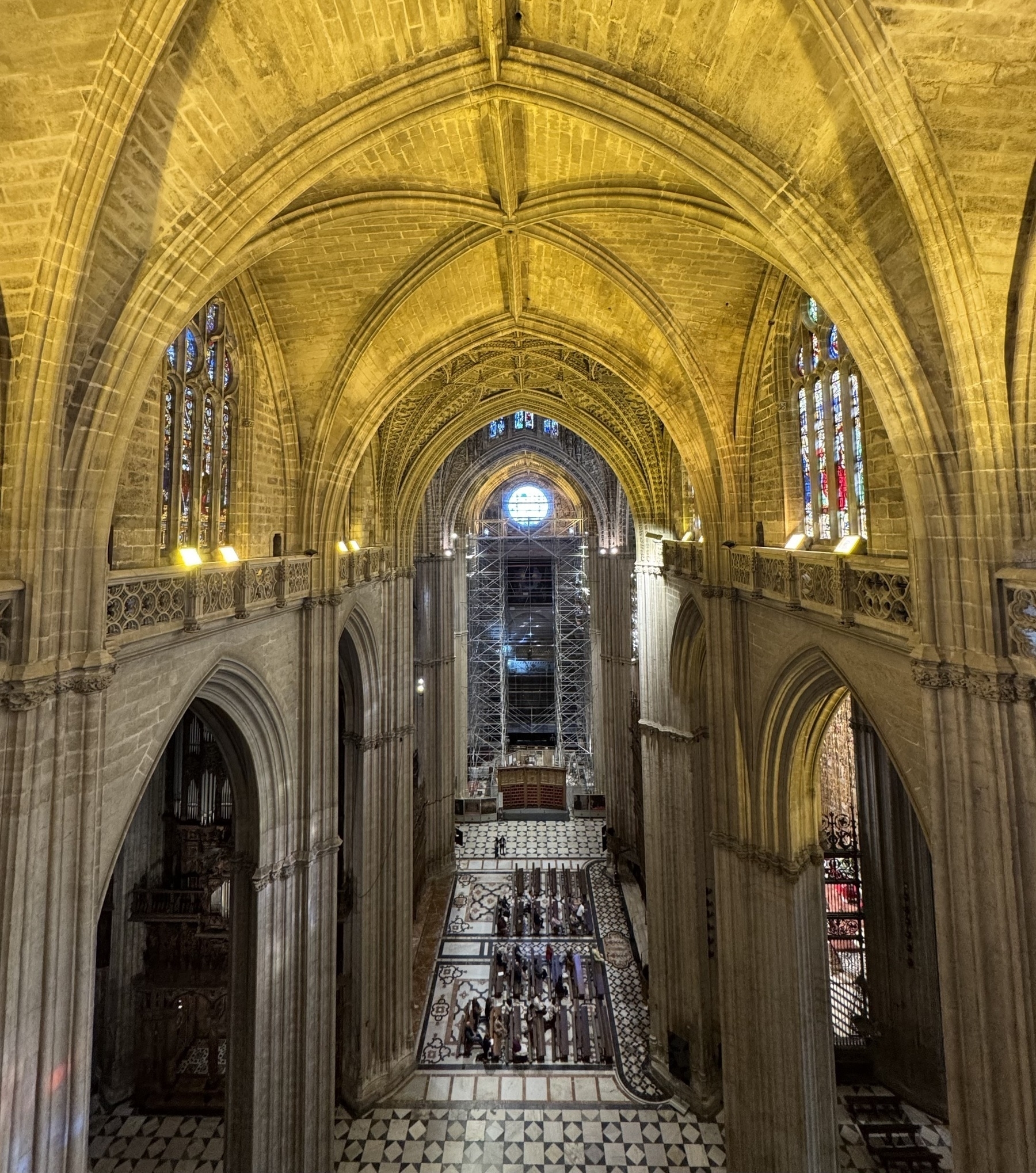 Sweeping view down the central nave from an elevated position, showcasing soaring ribbed vaults, arched columns, and patterned tile flooring below, with soft golden lighting accentuating the grandeur of the space.