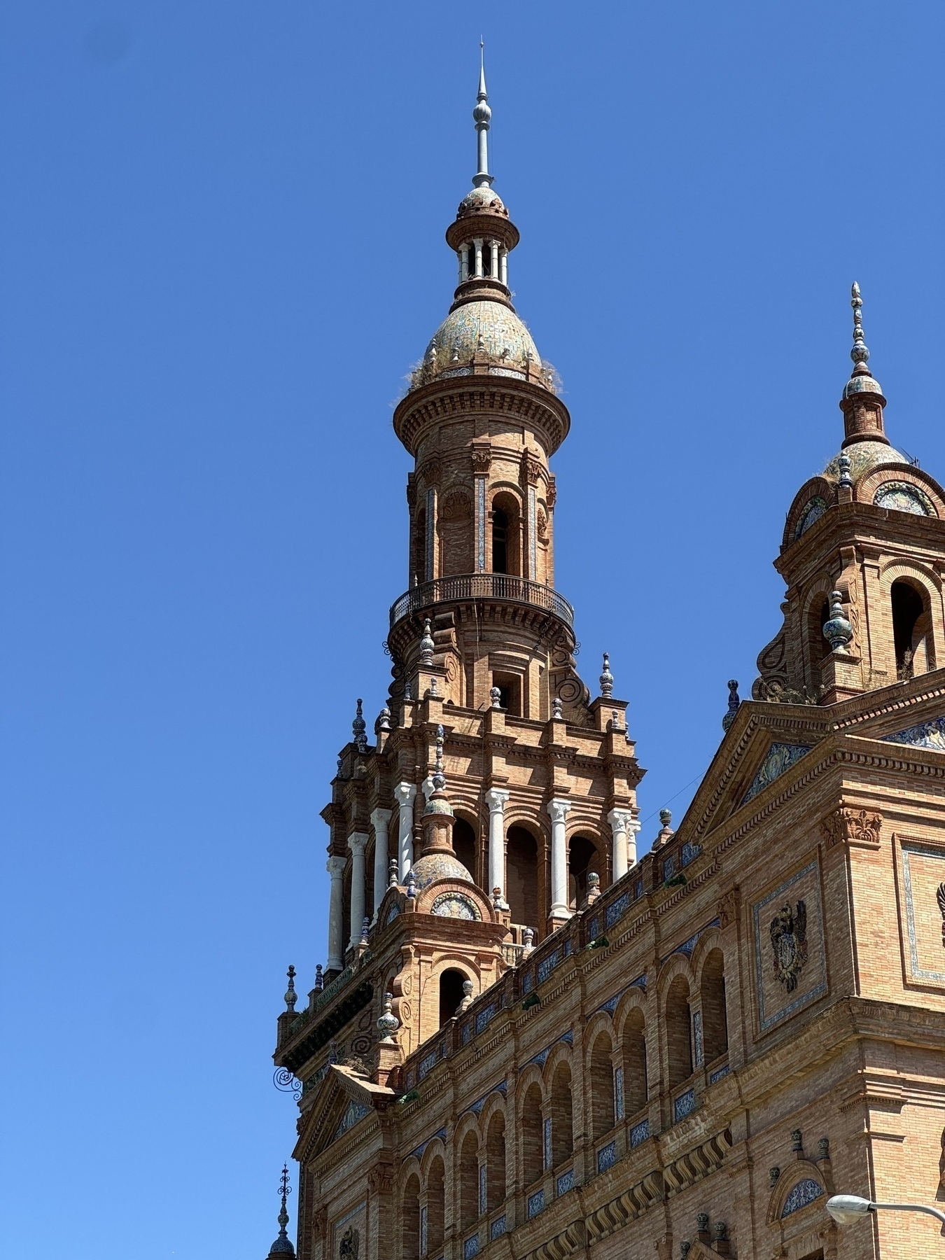 Close-up of the main tower at one end of the Plaza de España, showcasing intricate brickwork, decorative ceramic tile accents, arched openings, columns, and a domed top. The tower rises dramatically against a deep blue sky.