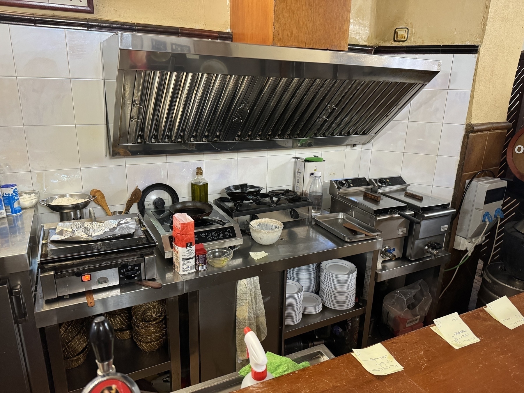 View of the restaurant’s open kitchen with a stainless steel countertop, stove, fryers, and shelves filled with plates and ingredients.